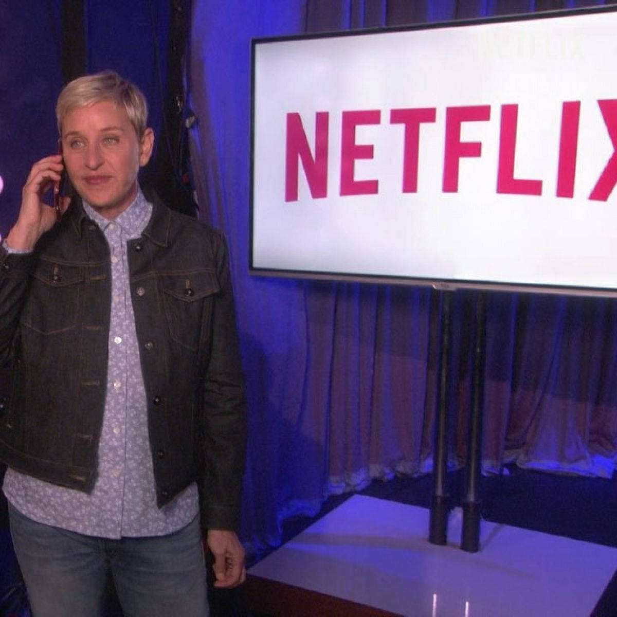 Netflix 桌布 壁紙 — 艾倫·狄珍妮在錄影棚內拿著電話，站在一個顯示Netflix標誌的大螢幕旁，舞台燈光為藍色。