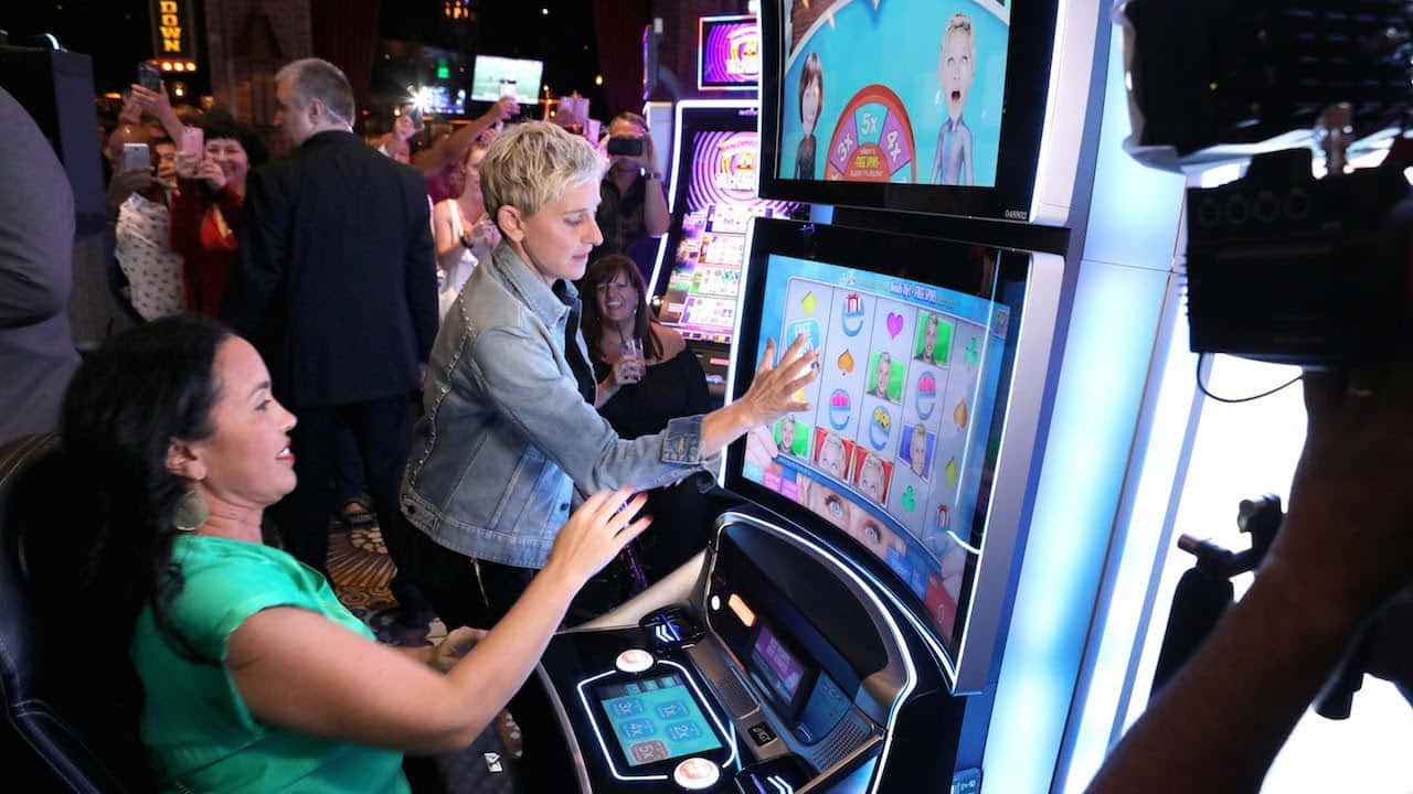 game 桌布 壁紙 — 艾倫·狄珍妮絲和一名女子在燈光明亮的賭場中玩色彩繽紛的吃角子老虎機遊戲，周圍有旁觀者和攝影機。