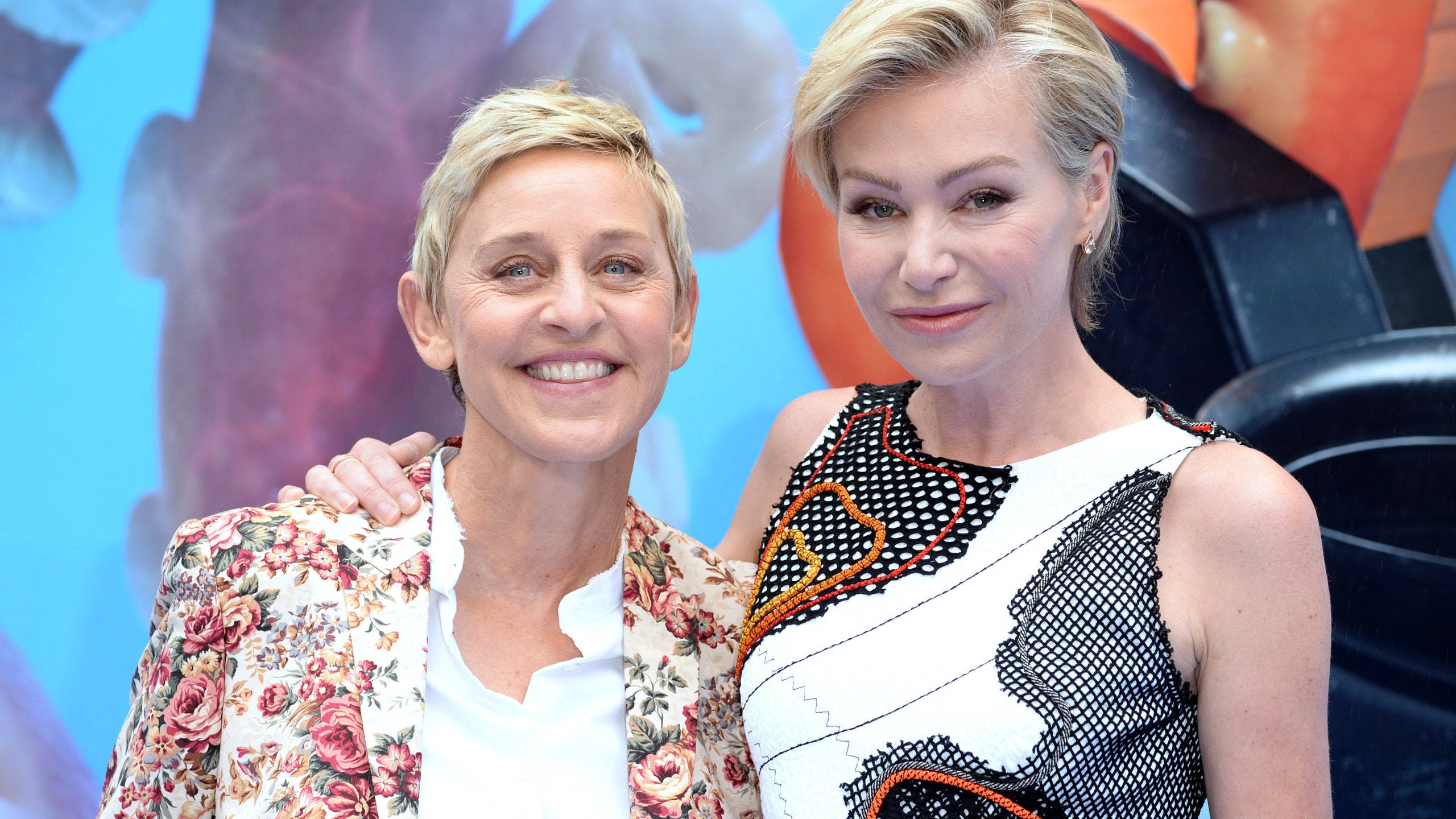 Ellen Degeneres 桌布 壁紙 — 艾倫·狄珍妮絲和波蒂亞·德羅西在色彩繽紛的抽象背景前，穿著鮮豔的花卉和幾何圖案服裝，對著鏡頭溫暖地微笑。