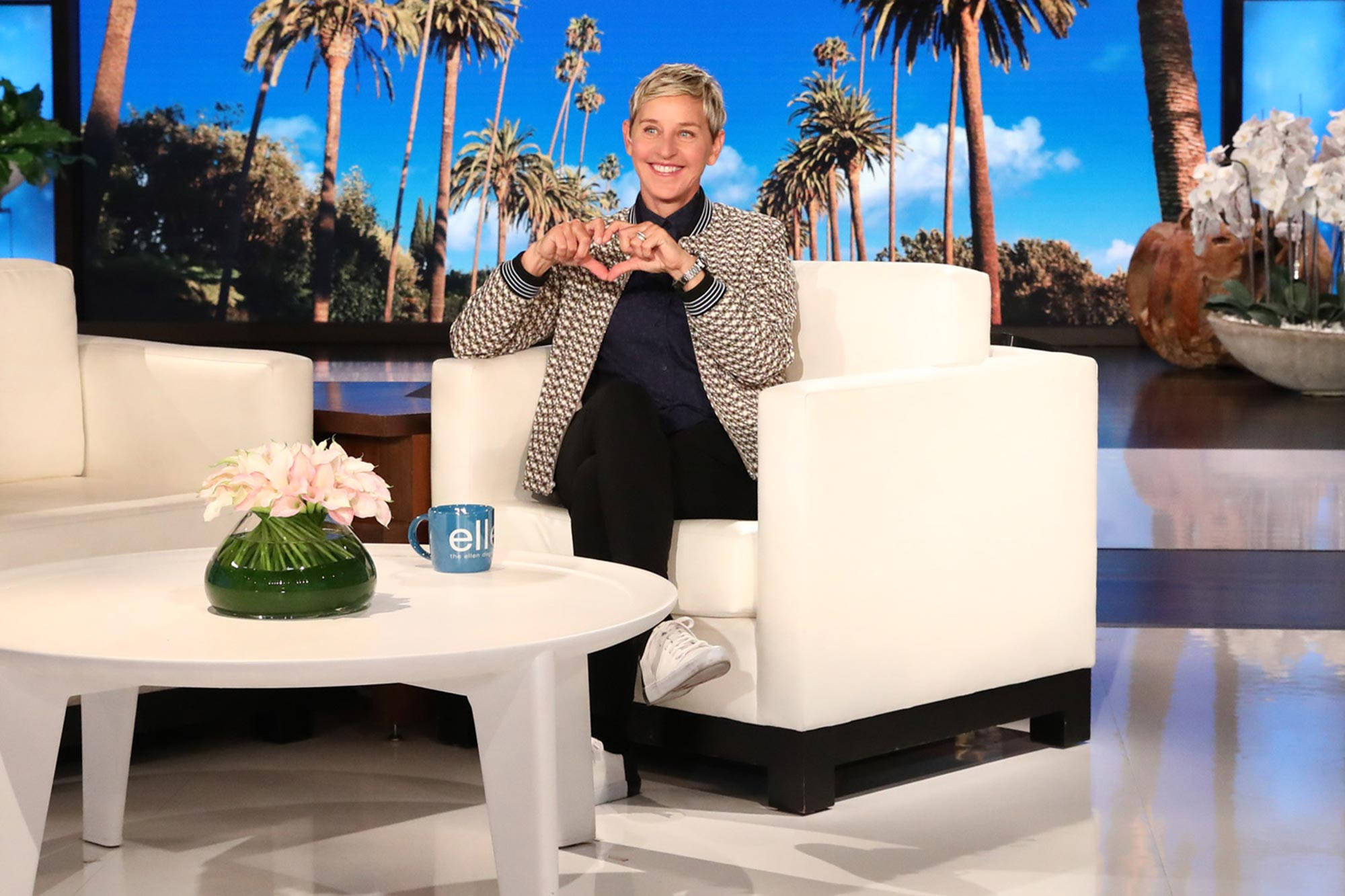 Ellen Degeneres 桌布 壁紙 — 艾倫·狄珍妮絲在棕櫚樹背景的白色場景中，用雙手比出愛心，露出溫暖的笑容。