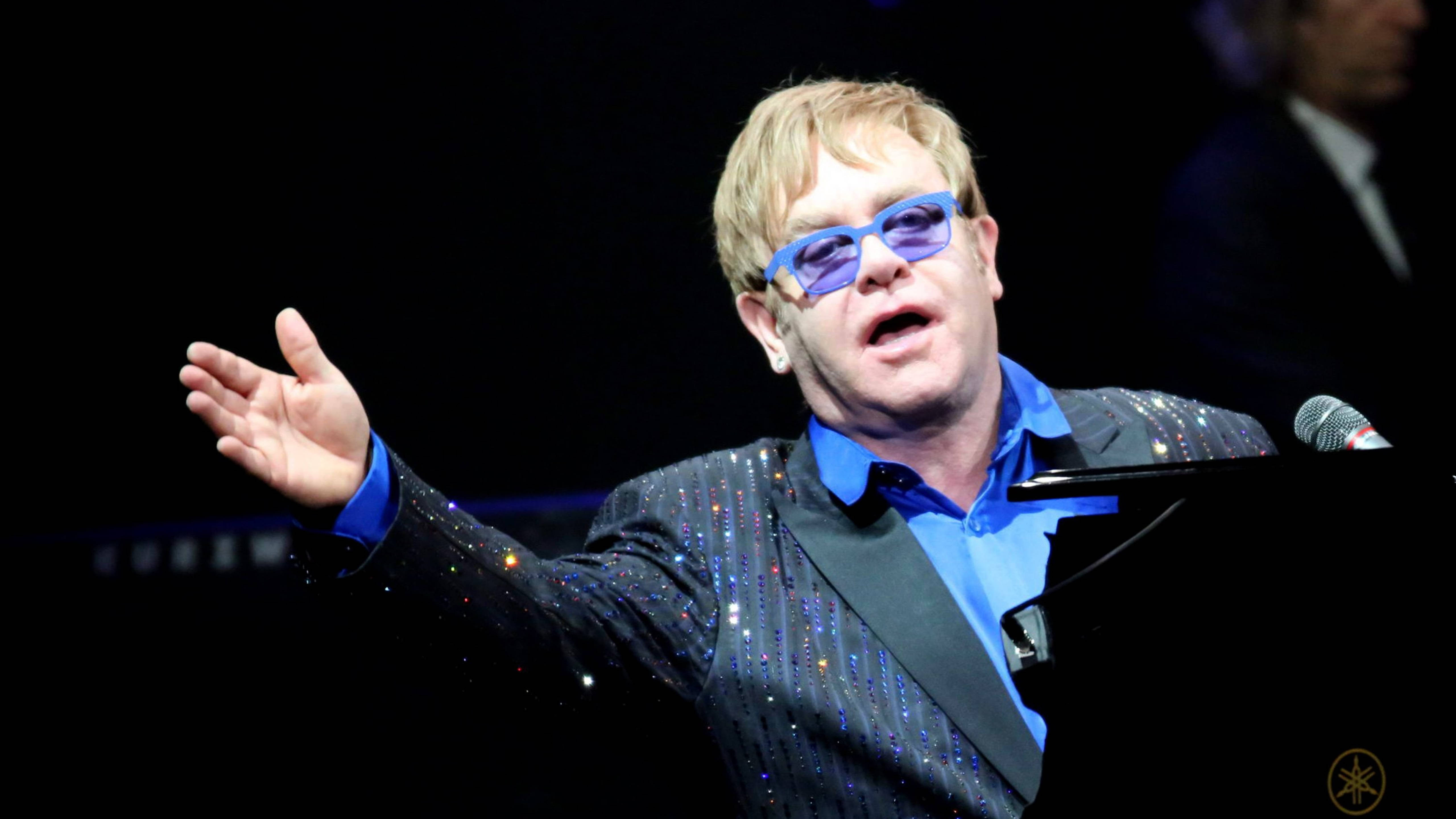 Elton John 桌布 壁紙 — 艾爾頓·強在舞台上表演，身穿閃亮黑色外套和藍色太陽眼鏡，一隻手臂伸展。