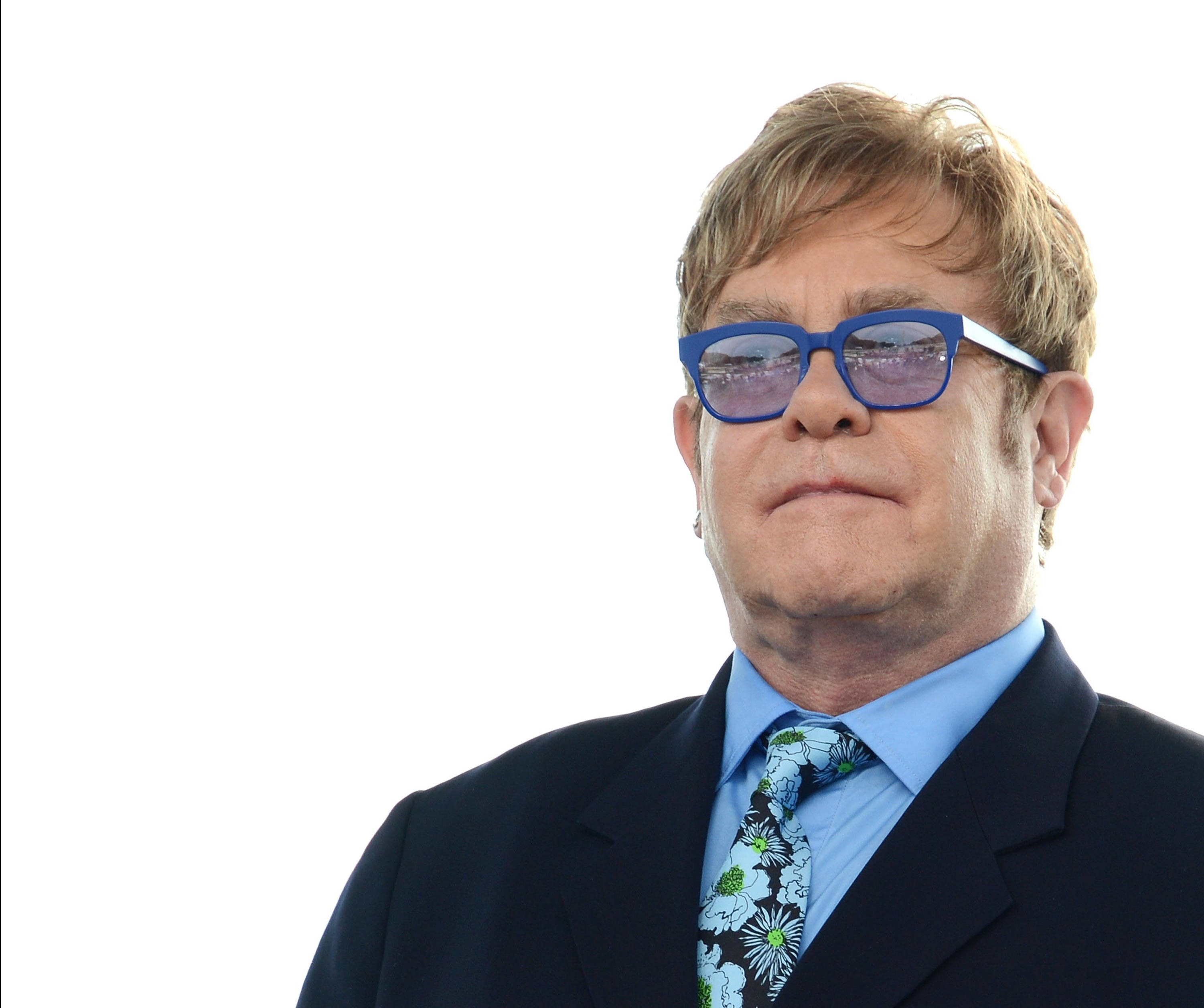 Elton John 桌布 壁紙 — 艾爾頓·強穿著深色西裝和花卉領帶，在純白背景下戴著大膽的藍色太陽眼鏡。