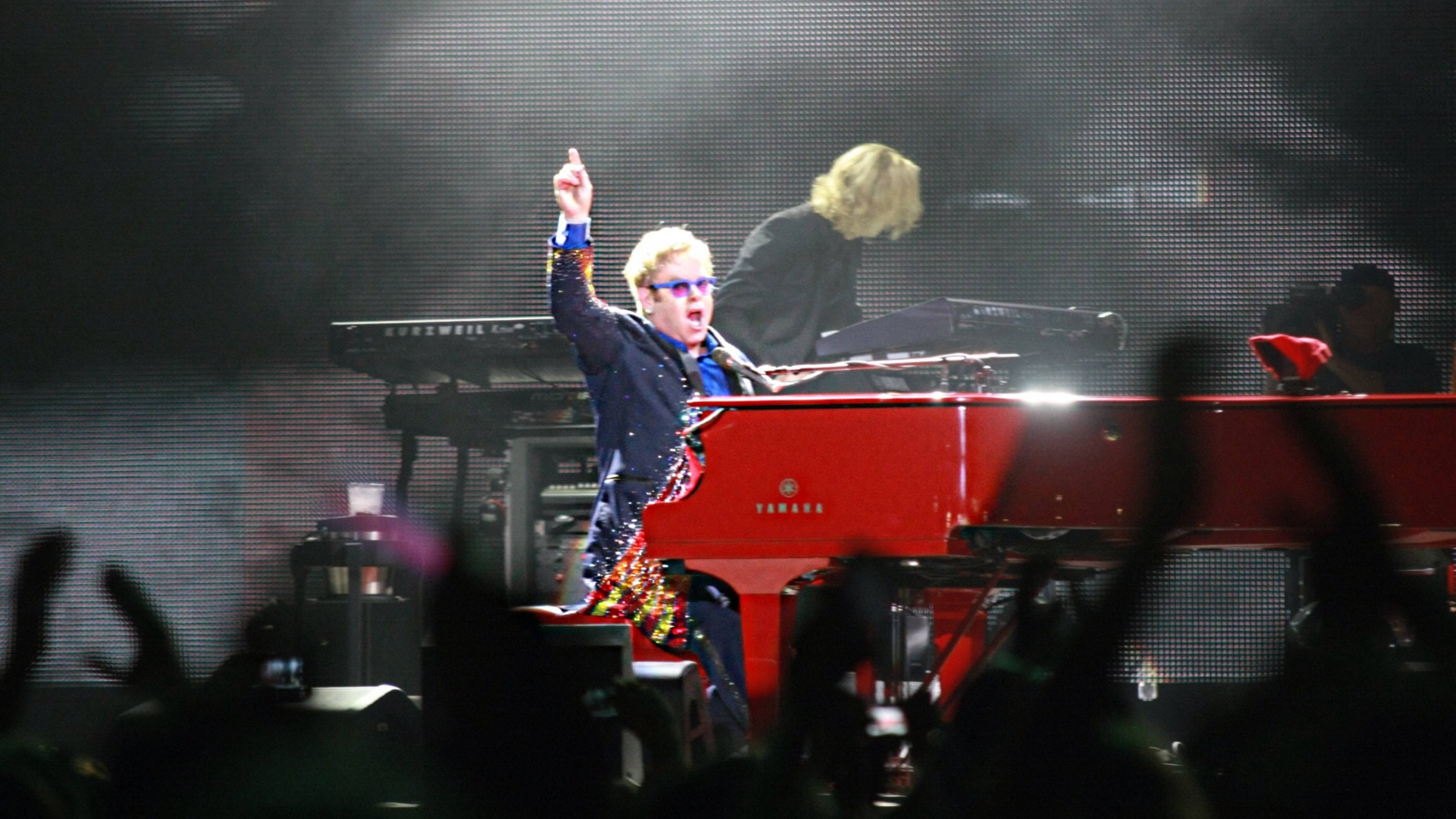 Elton John 桌布 壁紙 — 艾爾頓·約翰在紅色鋼琴上充滿活力地表演，在明亮的舞台燈光下，觀眾的手在前面舉起。
