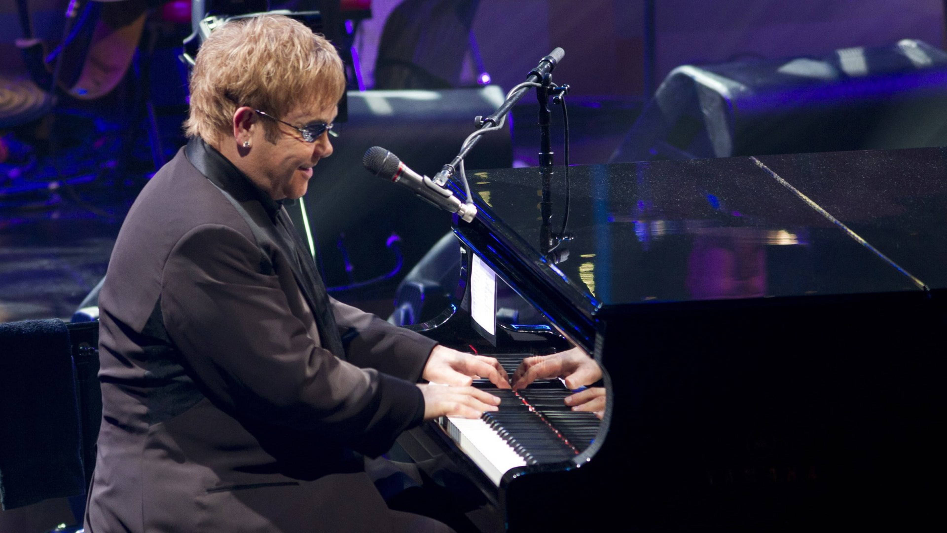 Elton John 桌布 壁紙 — 艾爾頓·約翰在紫色舞台燈光下專注地彈奏三角鋼琴。