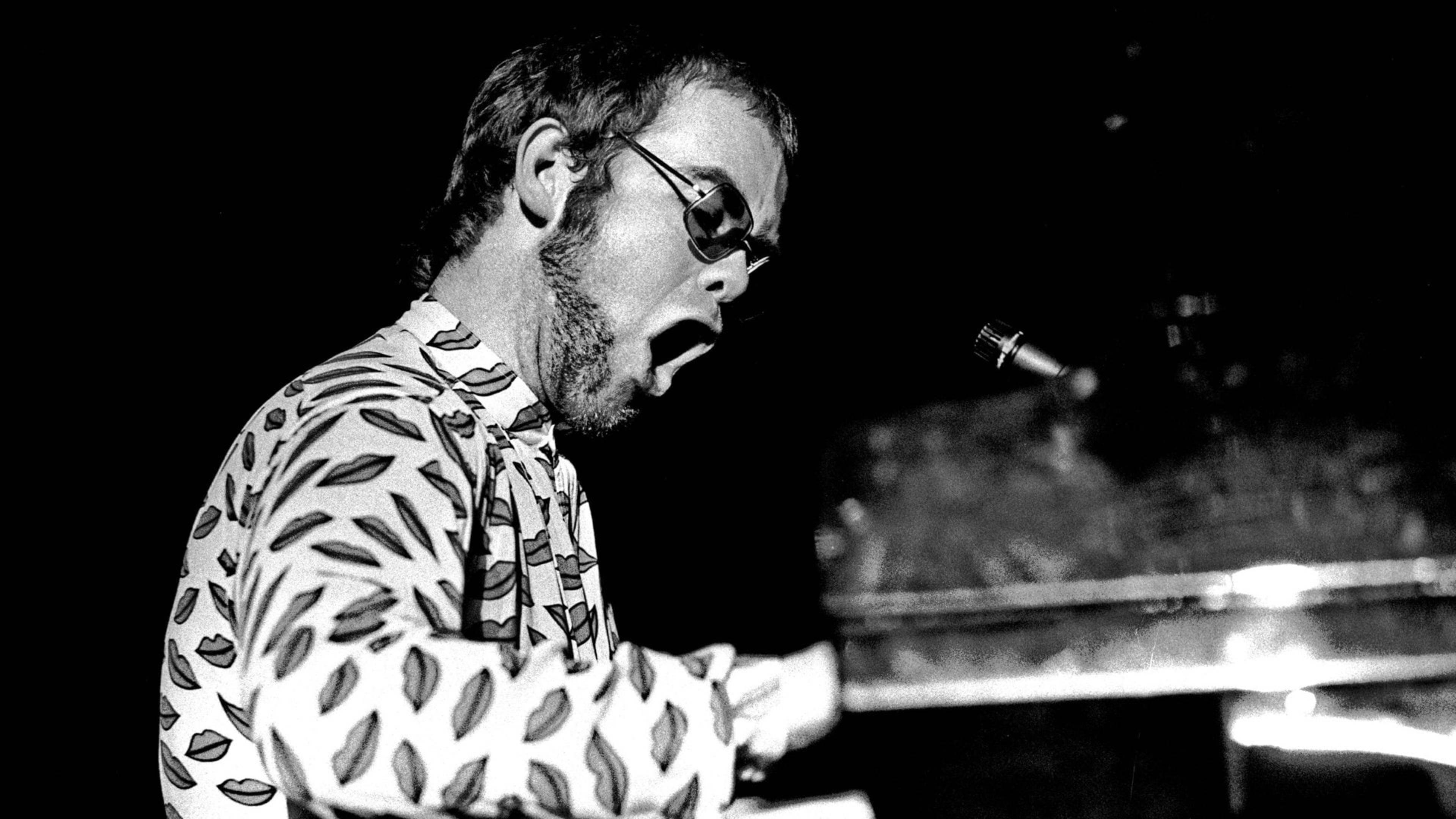 Elton John 桌布 壁紙 — 艾爾頓·約翰在舞台燈光下於鋼琴前熱情演唱的黑白照片。