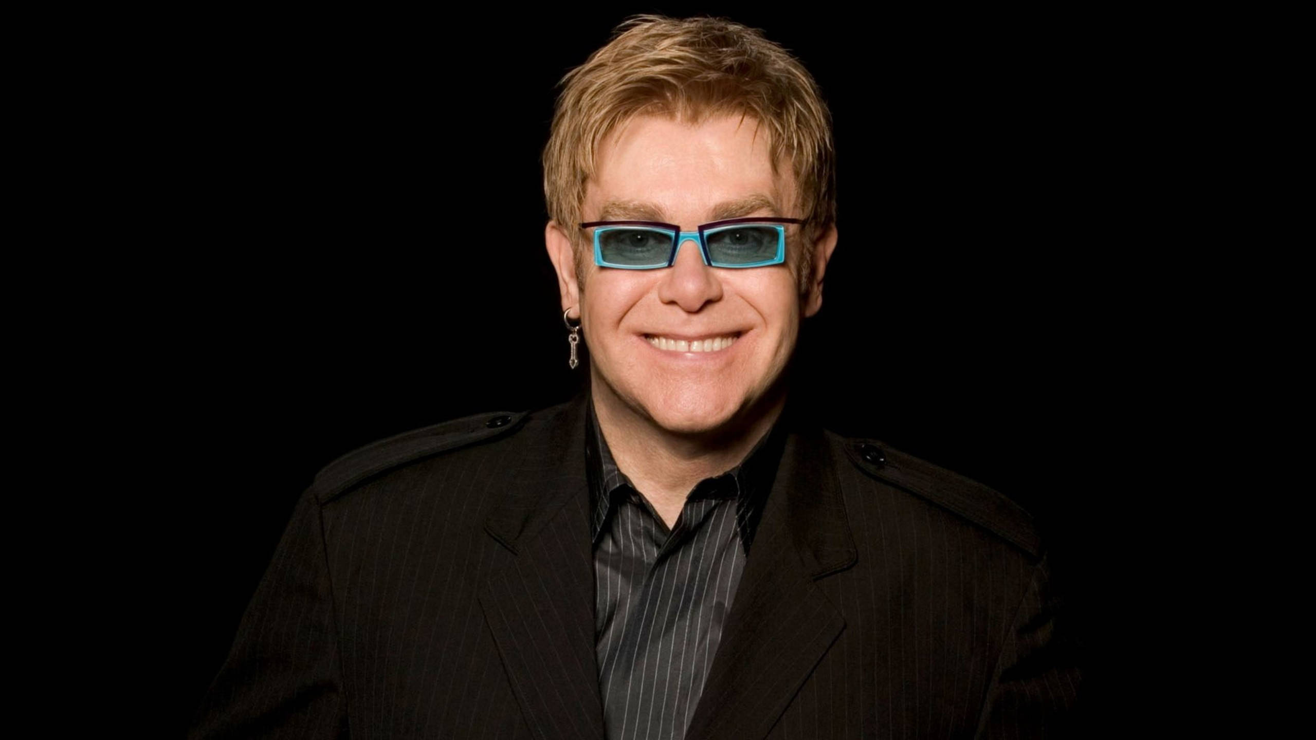 Elton John 桌布 壁紙 — 艾爾頓·約翰戴著藍色框眼鏡和黑色細條紋西裝，在純黑色背景前燦爛微笑。