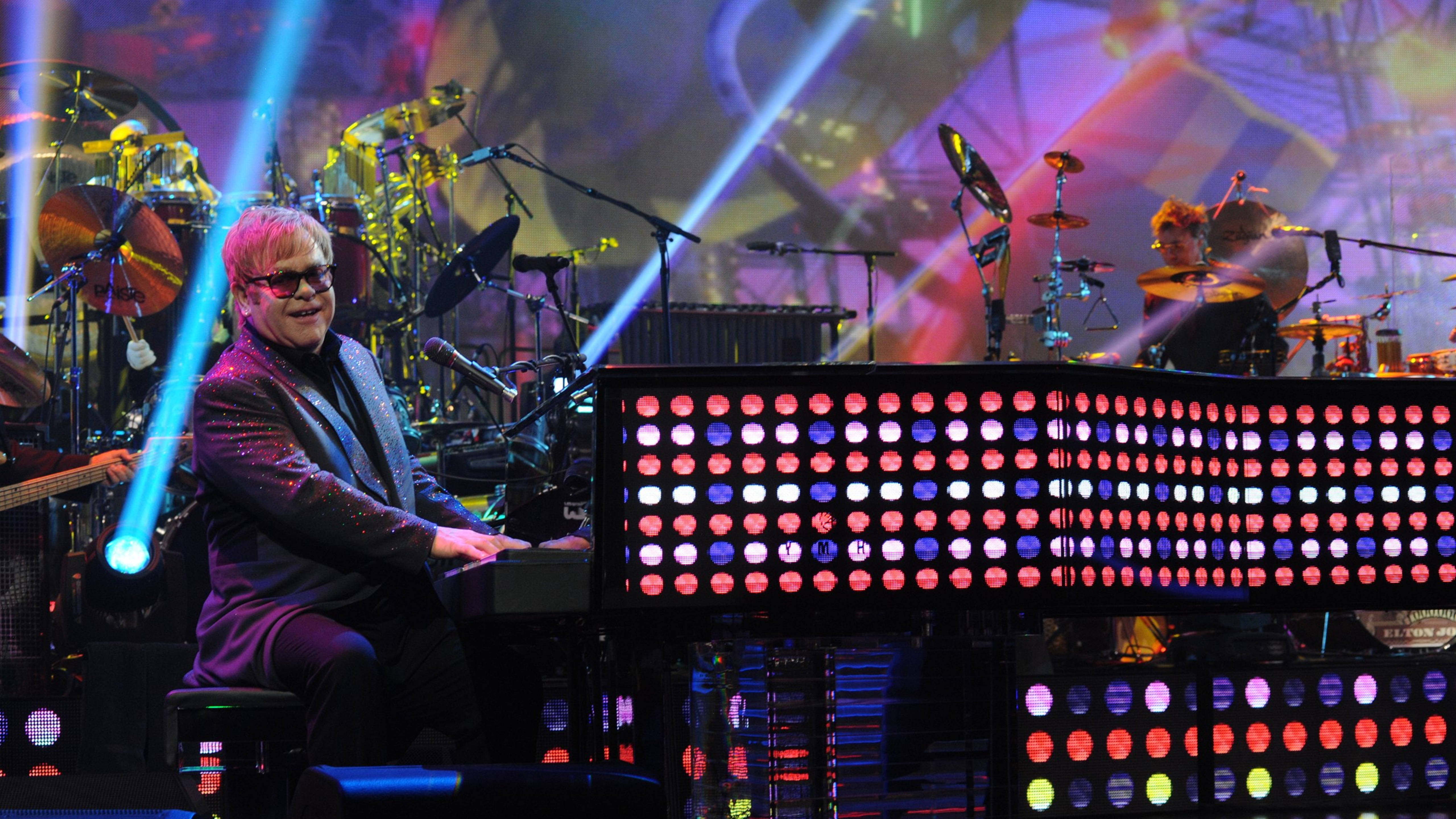 Elton John 桌布 壁紙 — 艾爾頓·約翰身穿閃亮西裝，在充滿活力的藍色聚光燈下表演，身後有色彩繽紛的LED鍵盤和鼓組。