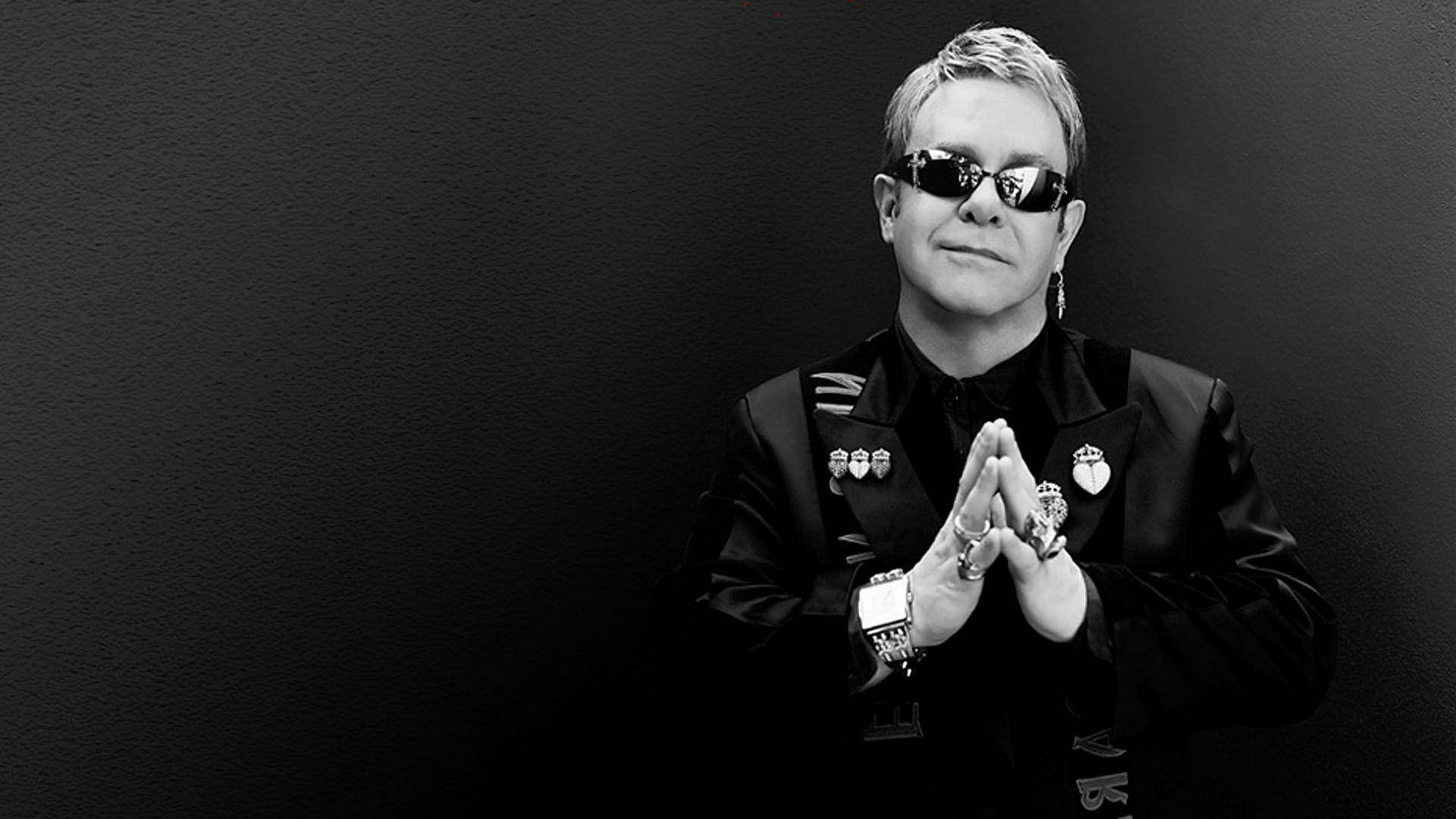 Rocketman 桌布 壁紙 — 艾爾頓強穿著黑色西裝、戴著太陽眼鏡，雙手合攏，在深色紋理背景前擺姿勢。