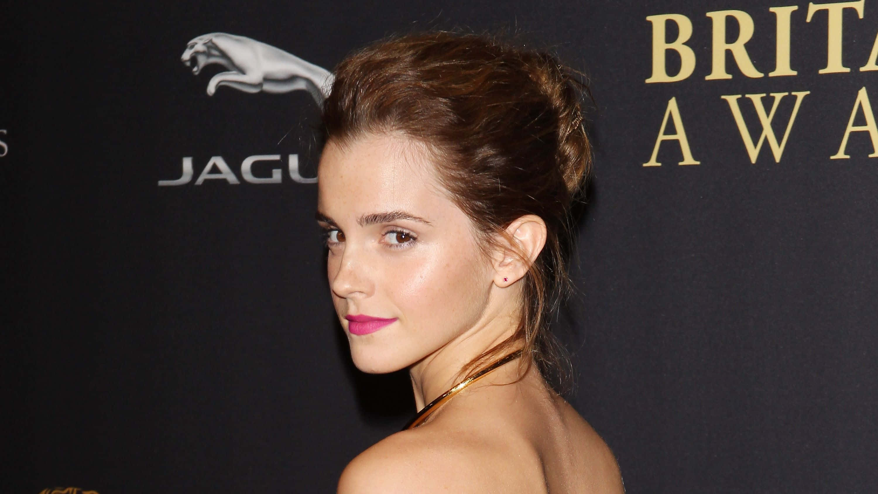Emma Watson 桌布 壁紙 — 艾瑪華森在Brit Awards頒獎典禮上，塗著粉紅色唇膏，戴著金色項鍊，在黑色背景前擺姿勢，背景上有Jaguar和Brit Awards標誌。