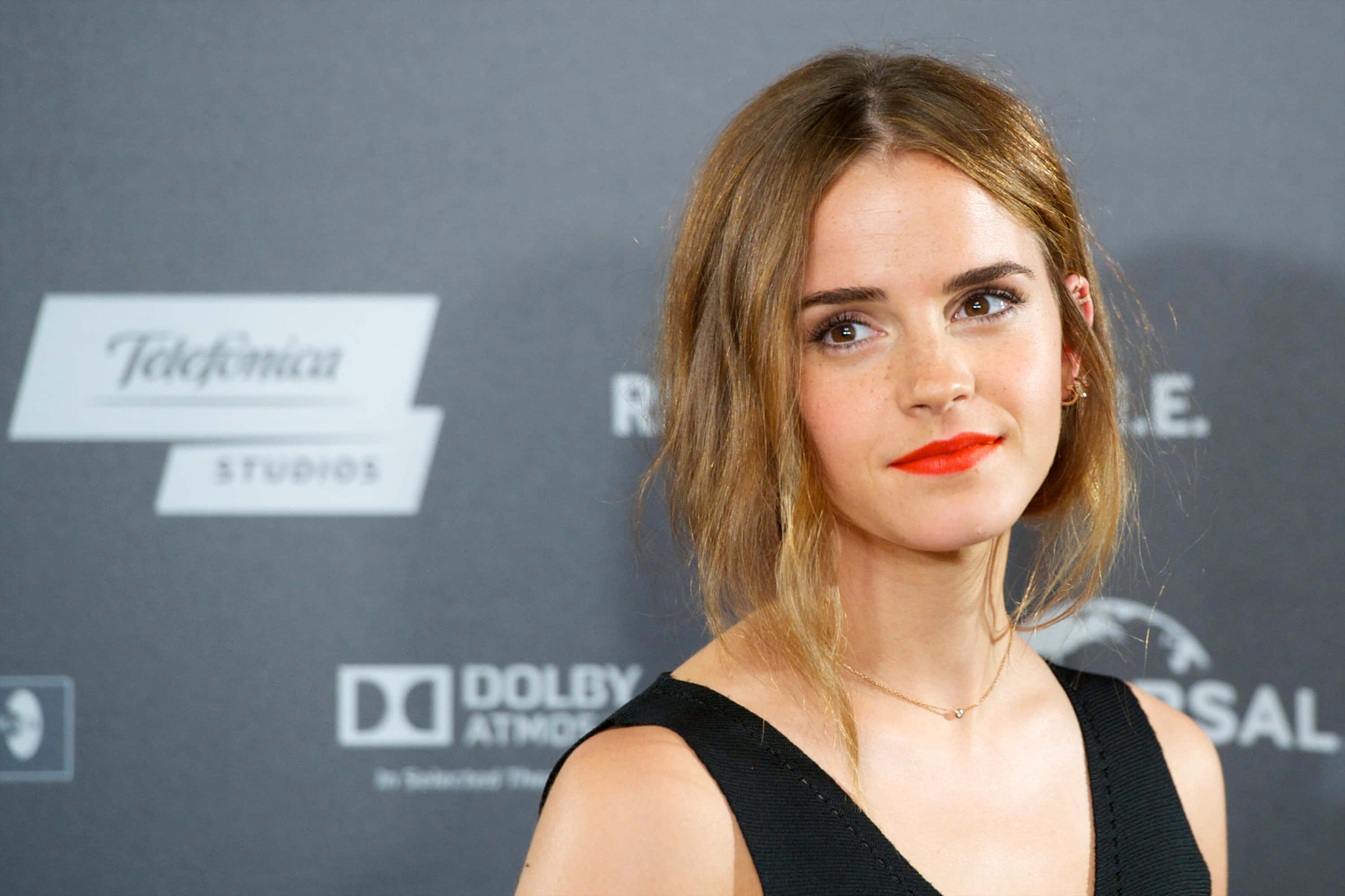 Emma Watson 桌布 壁紙 — 艾瑪華森在品牌活動背景前自信地擺姿勢，身穿黑色連身裙，搭配大膽的紅色唇膏。