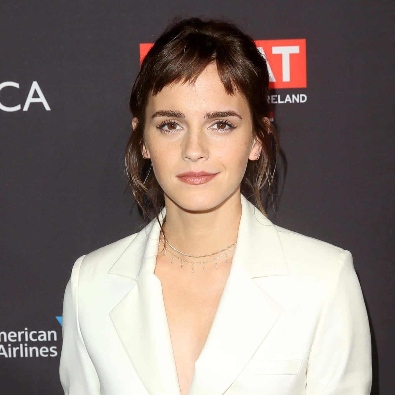 Emma Watson 桌布 壁紙 — 艾瑪華森在活動中穿著一件俐落的白色西裝外套，在深色品牌背景前擺姿勢。