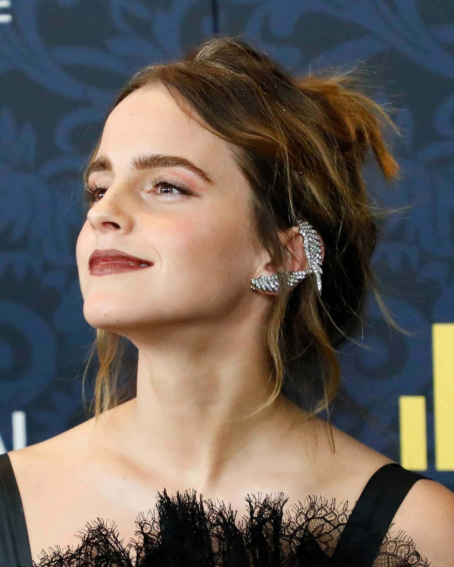 Emma Watson 桌布 壁紙 — 艾瑪華森抬頭微笑，戴著閃亮銀色耳環和黑色蕾絲上衣，背景為深藍色圖案。