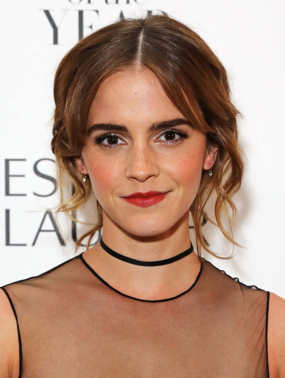 Emma Watson 桌布 壁紙 — 艾瑪華森穿著透視黑色上衣，塗上大膽紅唇，在白色活動背景前溫暖地微笑對著鏡頭。