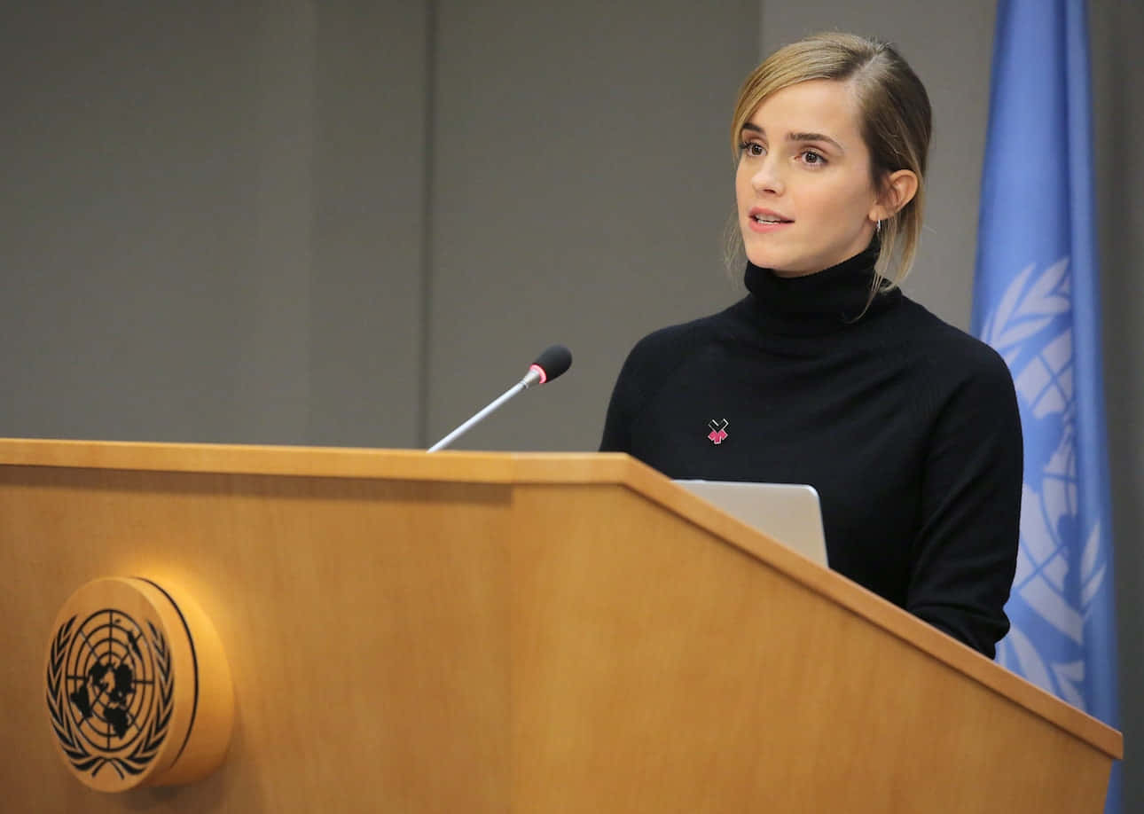 Emma Watson 桌布 壁紙 — 艾瑪華森穿著黑色高領毛衣，在藍色聯合國旗幟後方的木製聯合國講台上發言。