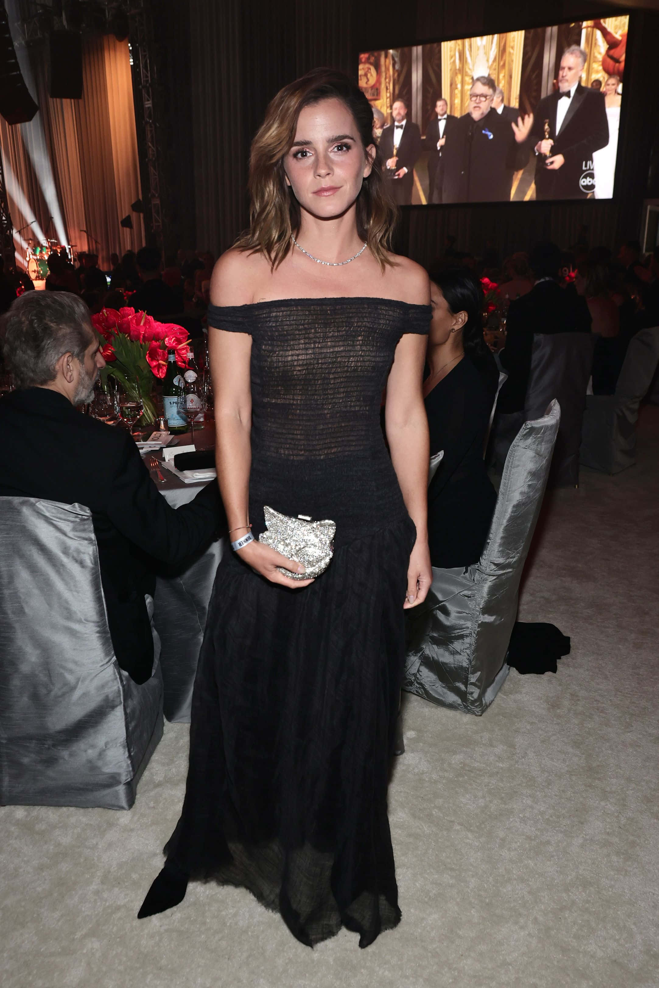 Emma Watson 桌布 壁紙 — 艾瑪華森身穿黑色露肩禮服，手持閃亮手拿包，在一個有賓客和大型螢幕的正式活動中優雅地站立。