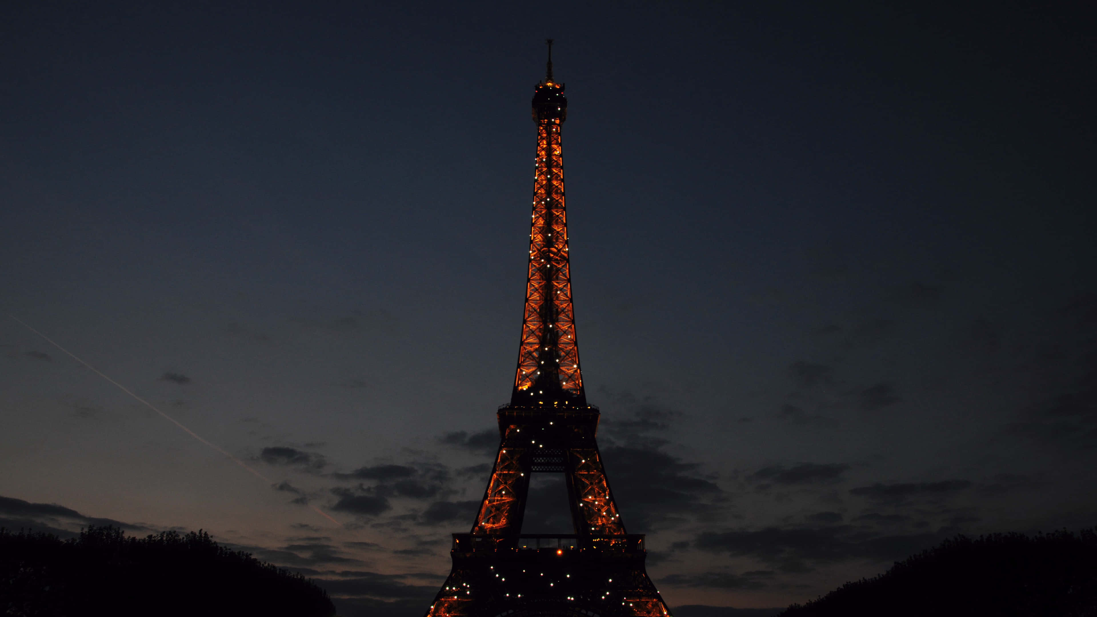 Paris At Night 桌布 壁紙 — 艾菲爾鐵塔在佈滿散雲和微弱飛機軌跡的暮色天空中溫暖地發光。