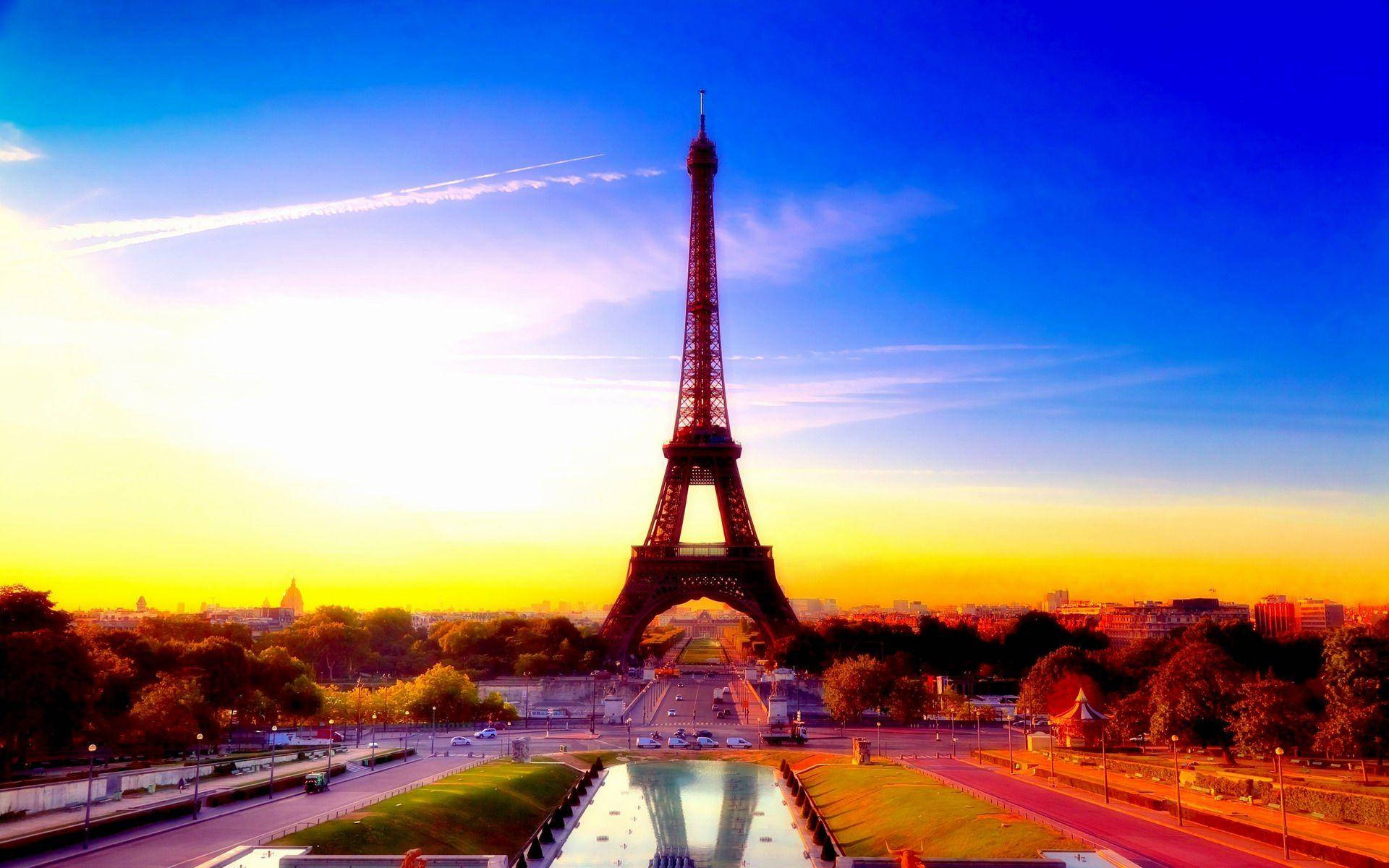 Paris Eiffel Tower 桌布 壁紙 — 艾菲爾鐵塔在充滿活力的日落天空下高聳矗立，金色的光芒照亮了巴黎的天際線，並在前景的水景中反射出來。