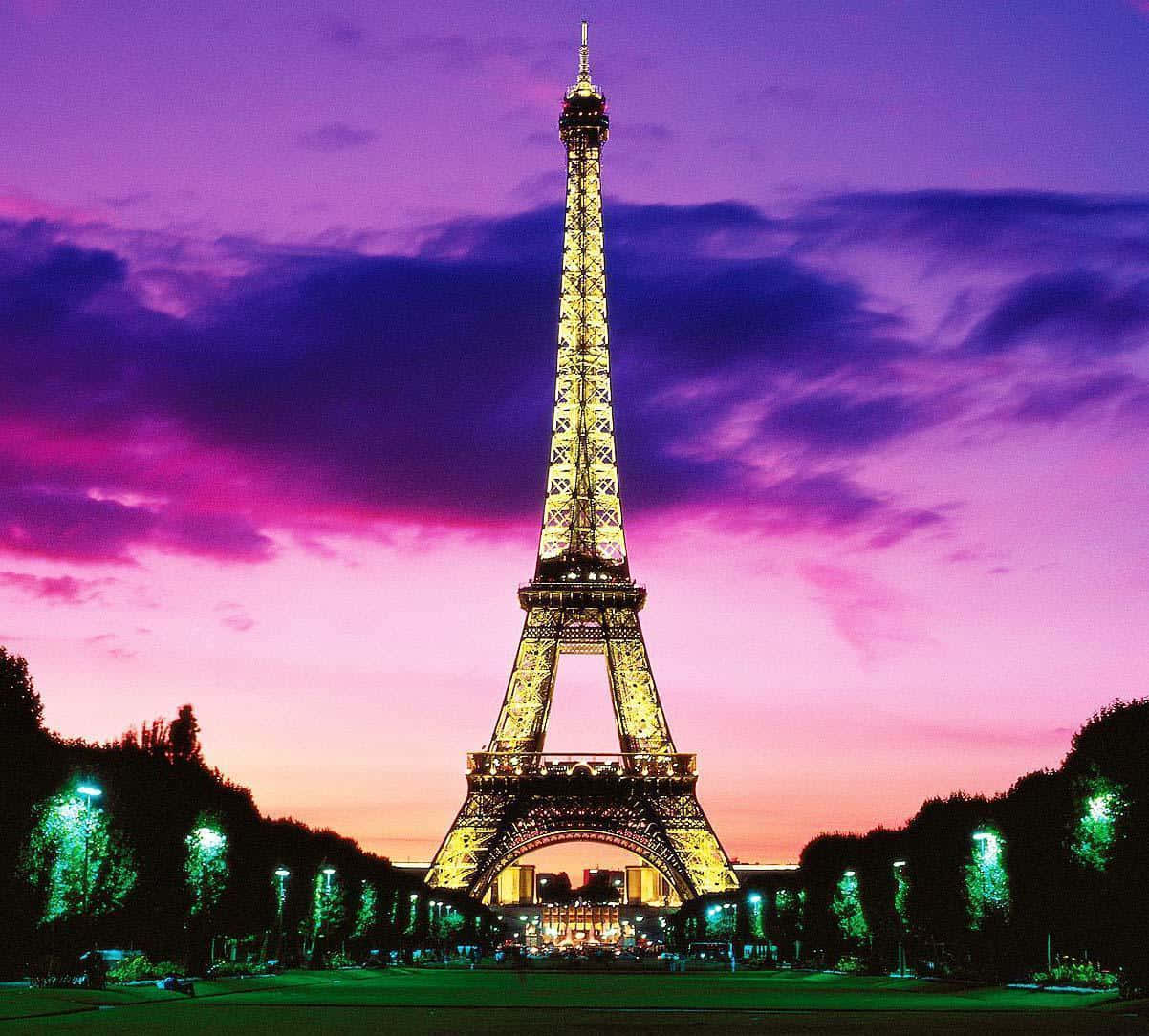 Eiffel Tower 桌布 壁紙 — 艾菲爾鐵塔在充滿活力的紫色和粉紅色暮色天空下閃耀著金色光芒，前景排列著燈光璀璨的樹木。