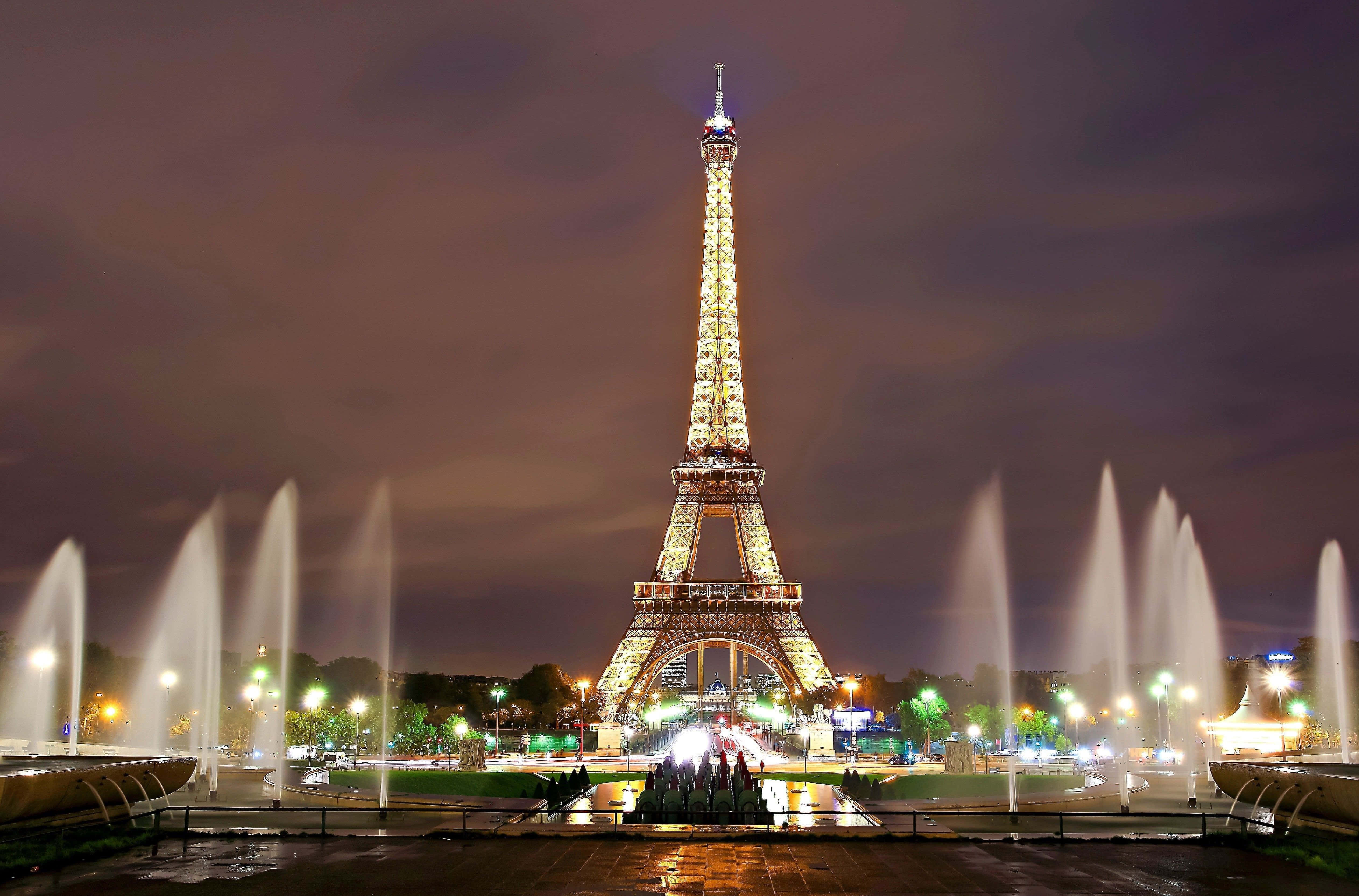 Paris Desktop 桌布 壁紙 — 艾菲爾鐵塔在夜晚燦爛發光，由對稱的噴泉在昏暗的天空下噴水構成框架。