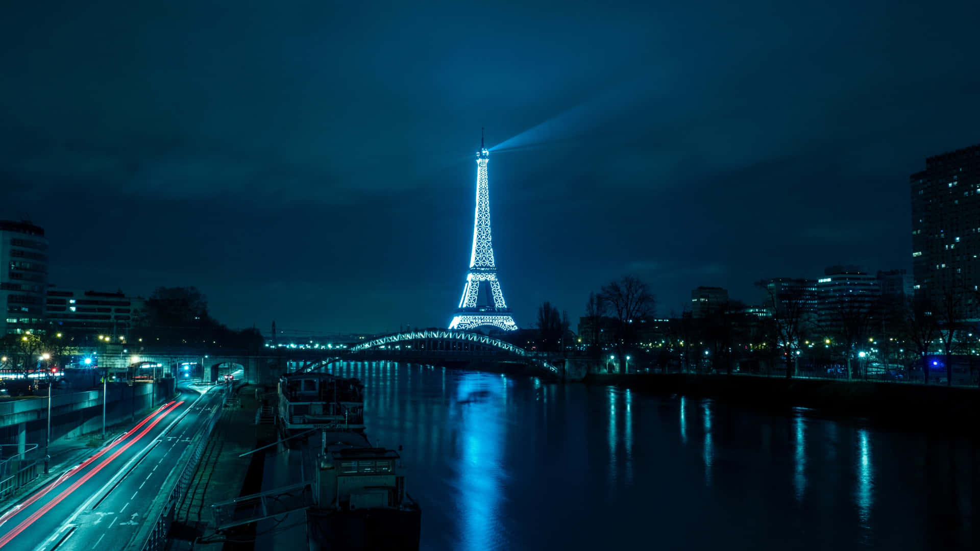 Eiffel Tower 桌布 壁紙 — 艾菲爾鐵塔在夜晚閃耀光芒，將光束投射到塞納河上，下方城市交通的燈光軌跡清晰可見。