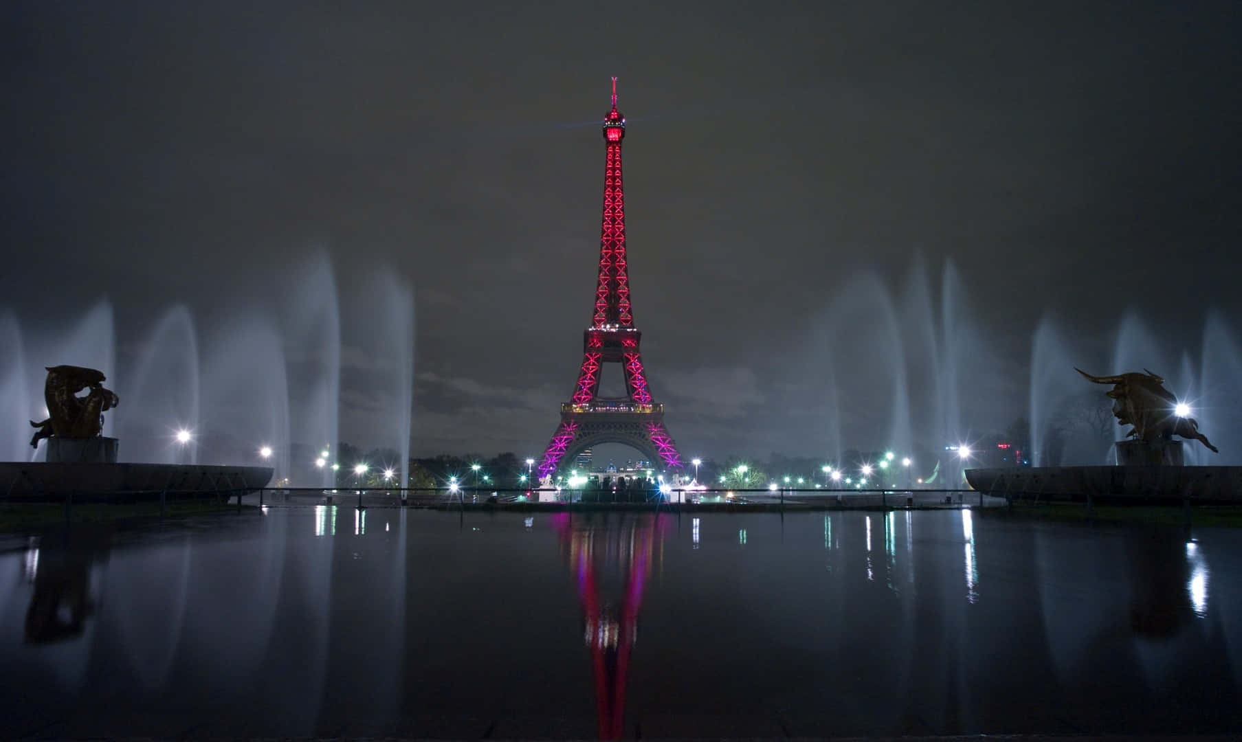 Eiffel Tower 桌布 壁紙 — 艾菲爾鐵塔在夜晚閃耀著粉紅色和紫色，倒映在平靜的池塘中，兩旁是燈光璀璨的噴泉和雕像。