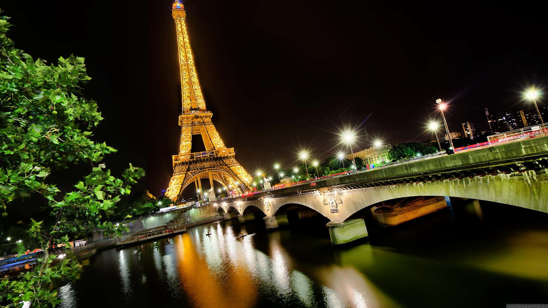 Eiffel Tower 桌布 壁紙 — 艾菲爾鐵塔在夜晚閃耀著金色光芒，燈光映照在石橋下的塞納河上，左側有茂密的綠樹作為框架。