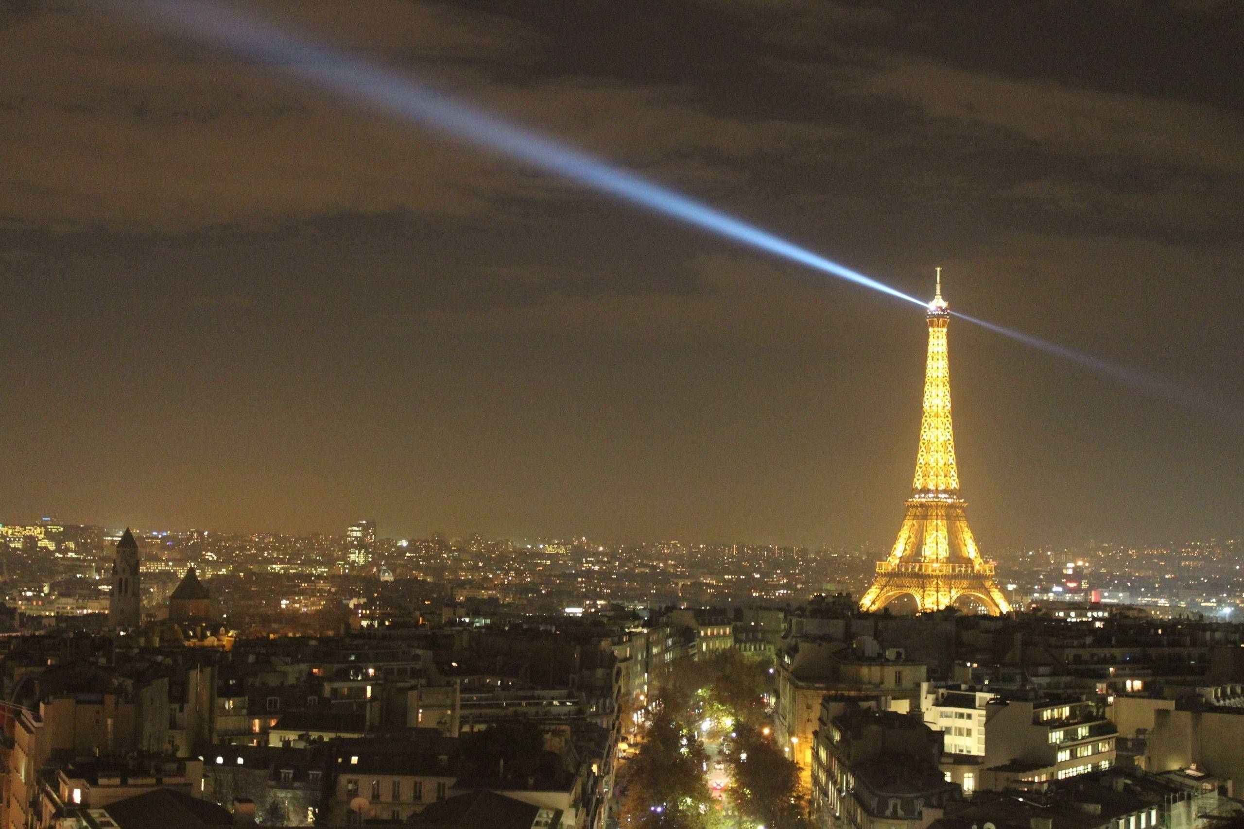 Paris Eiffel Tower 桌布 壁紙 — 艾菲爾鐵塔在夜晚閃耀著金色光芒，一道強勁的藍色雷射光束劃過巴黎上空多雲的天空。