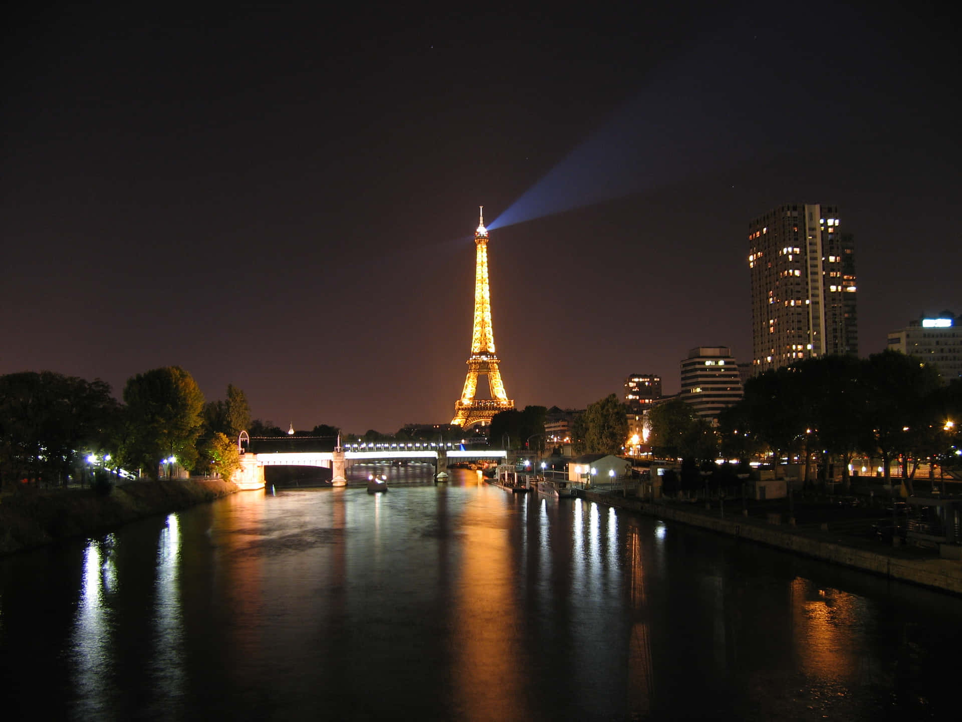 Eiffel Tower 桌布 壁紙 — 艾菲爾鐵塔在夜晚閃耀金色光芒，將光束射入塞納河上方的黑暗天空，城市燈光倒映在水面上。
