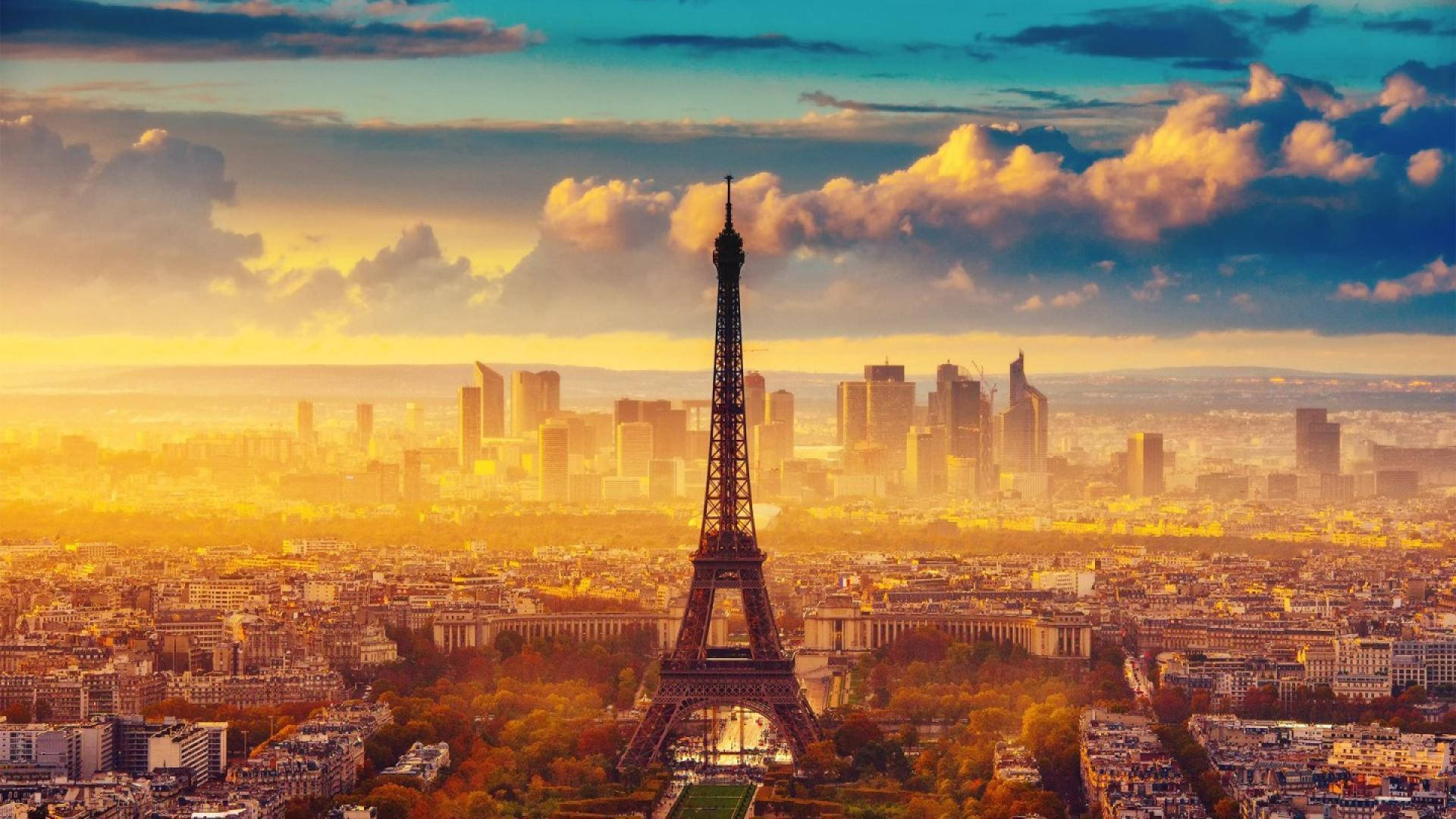 Eiffel Tower 桌布 壁紙 — 艾菲爾鐵塔在巴黎屋頂和現代摩天大樓之上，襯著金色夕陽天空和戲劇性的雲朵高高聳立。
