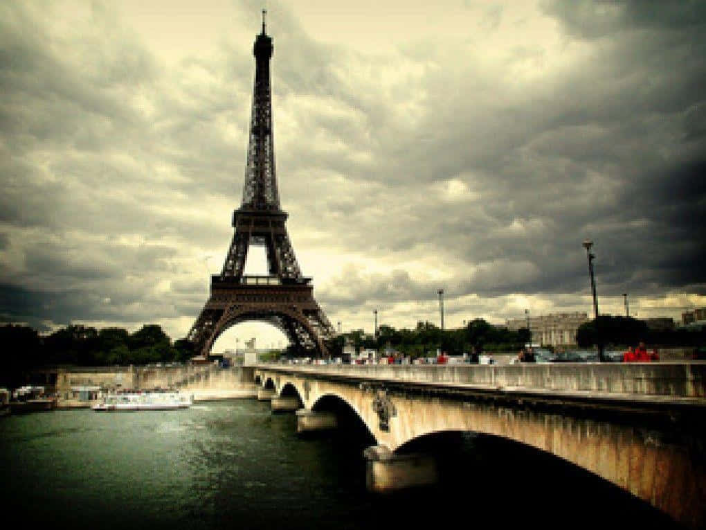 Eiffel Tower 桌布 壁紙 — 艾菲爾鐵塔在戲劇性的多雲天空下巍然聳立，橋樑和河船框住了這個標誌性的巴黎地標。