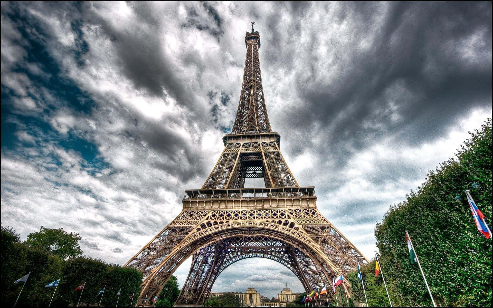 Eiffel Tower 桌布 壁紙 — 艾菲爾鐵塔在戲劇性的多雲天空下高聳矗立，底部環繞著茂密的綠樹和五彩繽紛的旗幟。