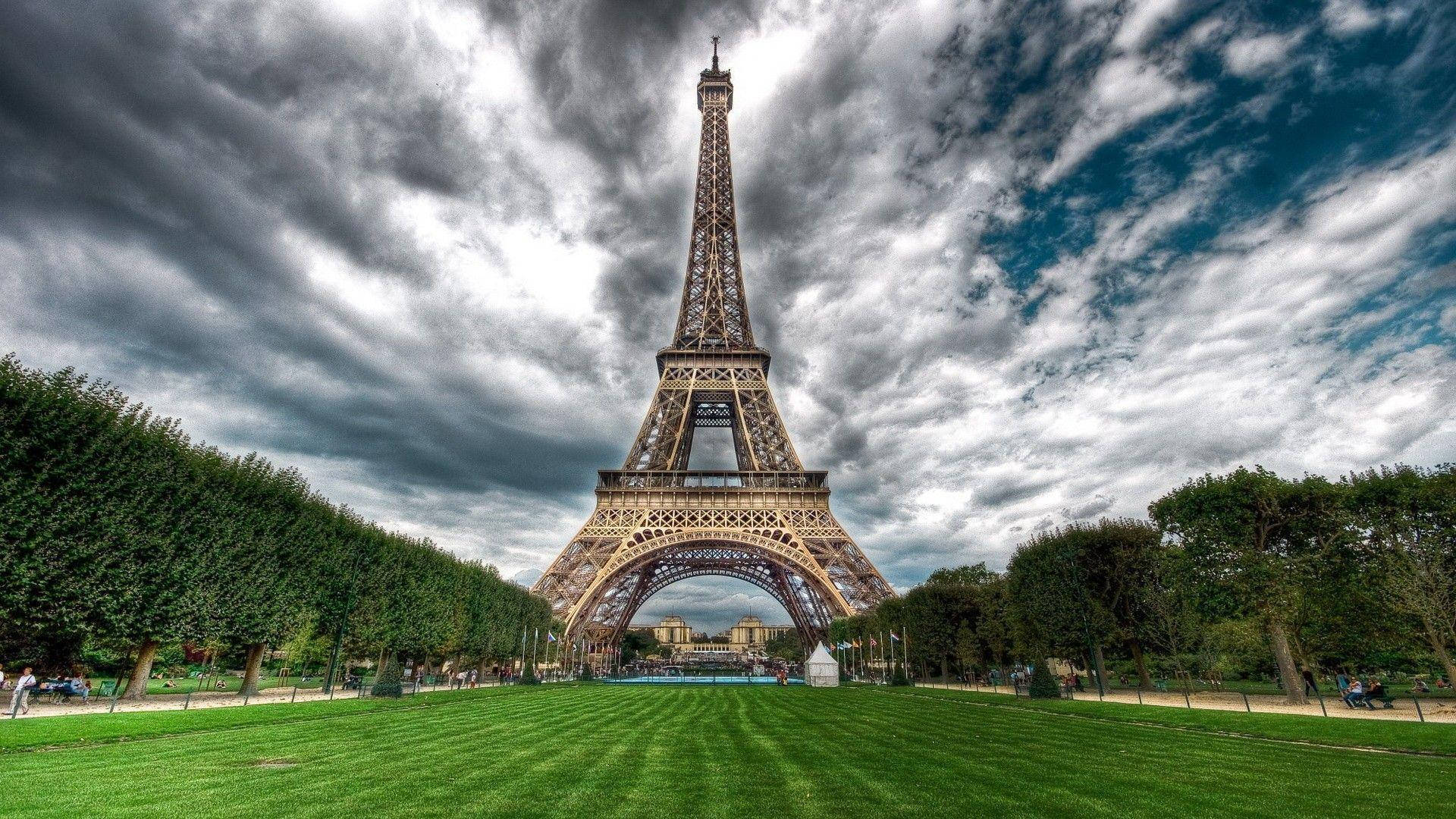 Eiffel Tower 桌布 壁紙 — 艾菲爾鐵塔在戲劇性的多雲天空下高高聳立，前景有充滿活力的綠色草坪和修剪整齊的樹木。