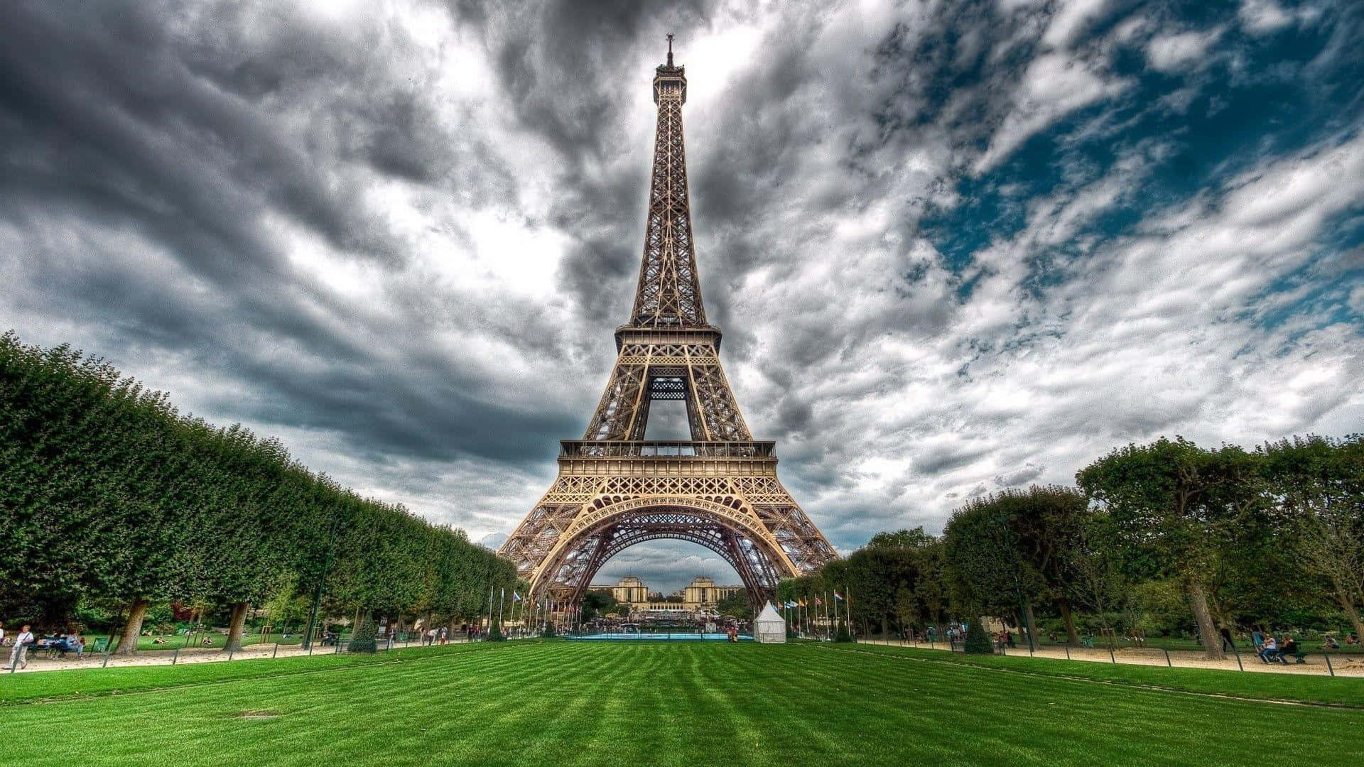 Eiffel Tower 桌布 壁紙 — 艾菲爾鐵塔在戲劇性的多雲天空下高高聳立，充滿活力的綠色草坪和修剪整齊的樹木襯托著這個標誌性的巴黎地標。