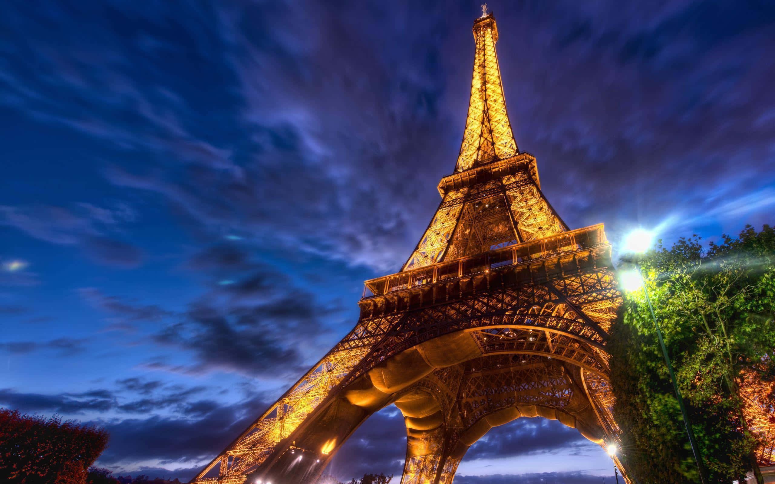 Eiffel Tower 桌布 壁紙 — 艾菲爾鐵塔在戲劇性的暮色天空下溫暖地發光，天空中有旋轉的雲朵和鮮豔的藍色色調。
