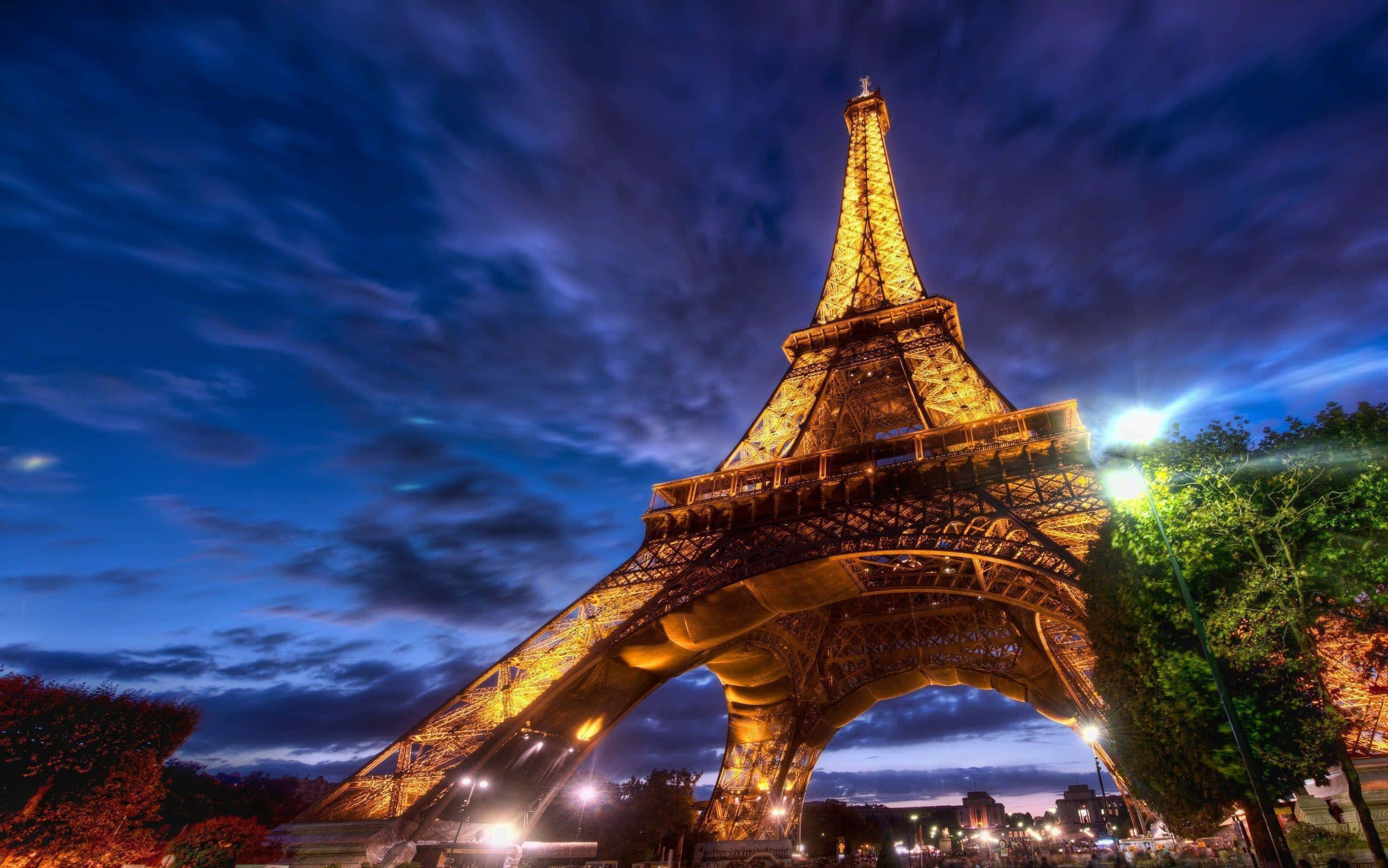 Eiffel Tower 桌布 壁紙 — 艾菲爾鐵塔在戲劇性的暮色天空下溫暖地發光，天空中有旋轉的雲彩，下方城市燈光閃爍。