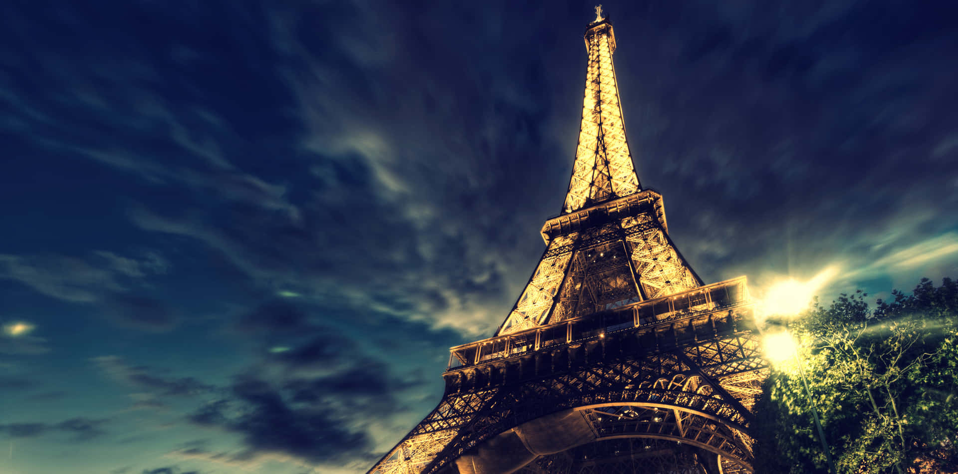 Eiffel Tower 桌布 壁紙 — 艾菲爾鐵塔在戲劇性的暮色天空中，伴隨著旋轉的雲彩和金色光線的照耀下亮起。