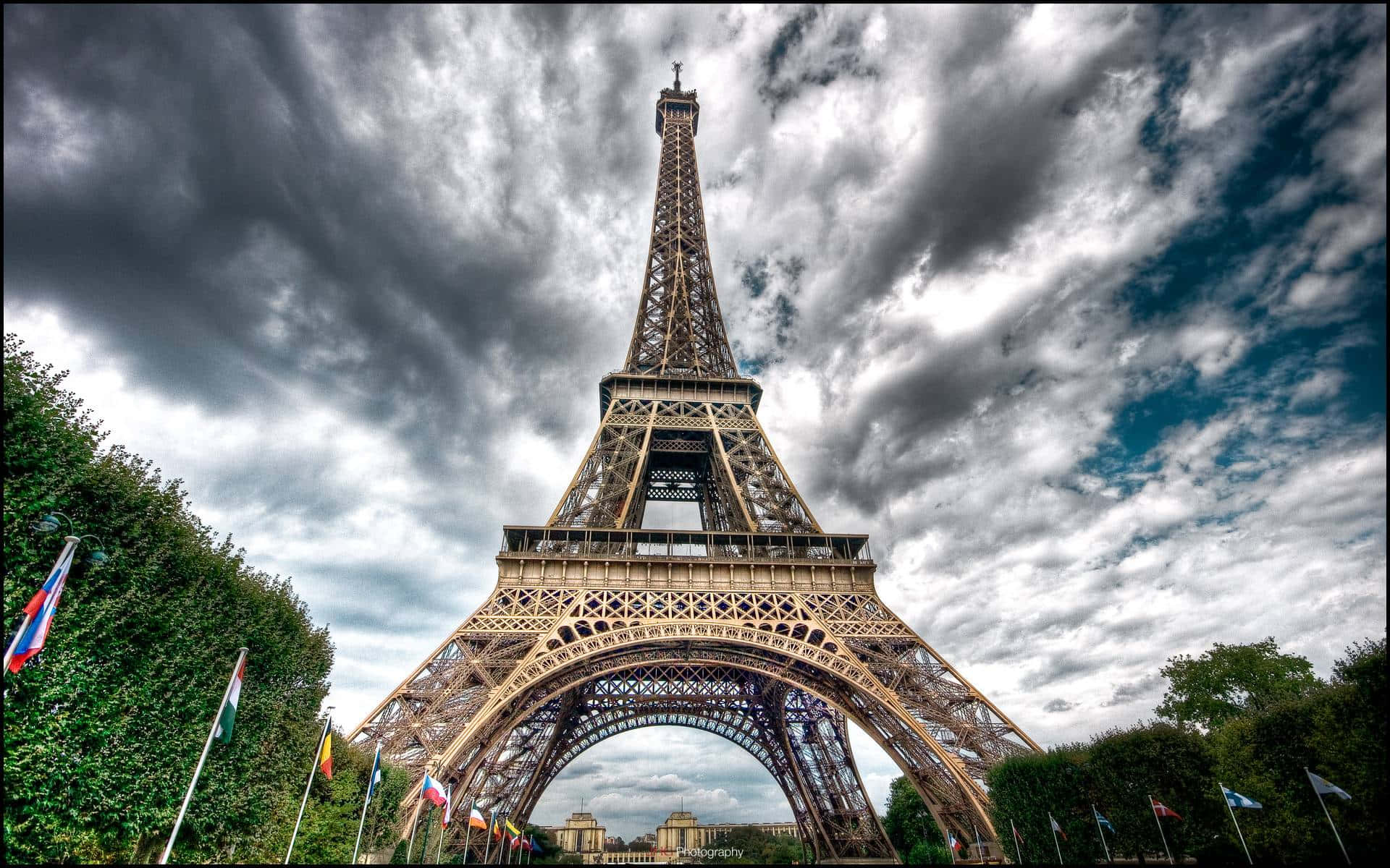 Eiffel Tower 桌布 壁紙 — 艾菲爾鐵塔在戲劇性的暴風雲下高聳矗立，底部有茂密的綠色樹籬和色彩繽紛的旗幟排列。