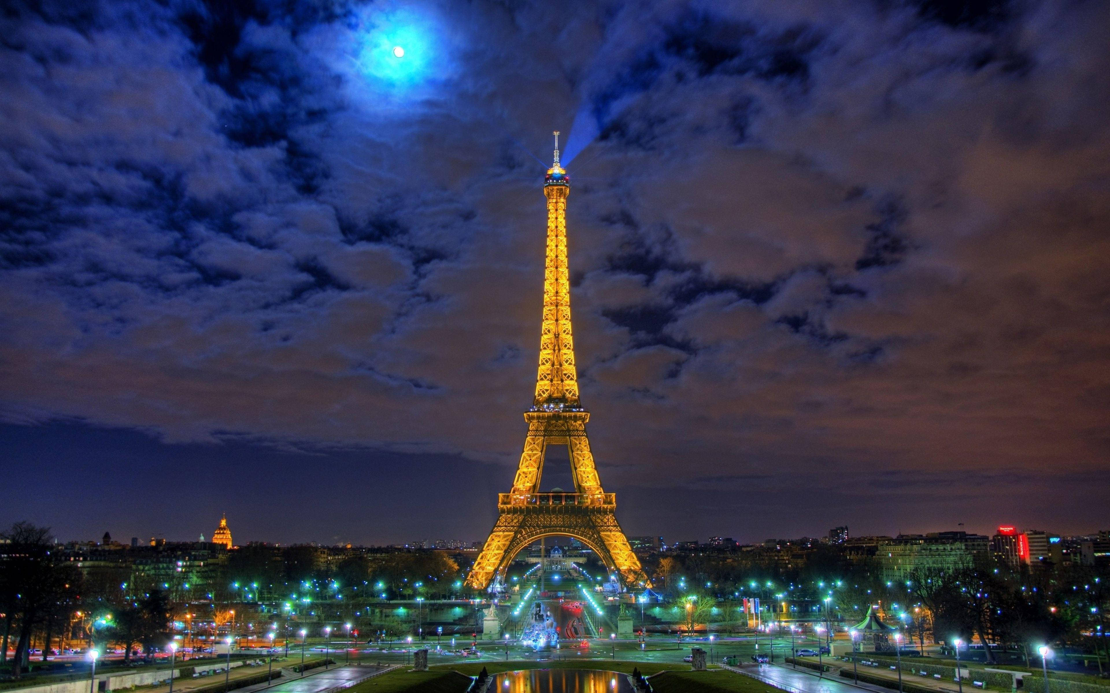 Paris Eiffel Tower 桌布 壁紙 — 艾菲爾鐵塔在戲劇性的藍色月光天空下閃耀著金色光芒，下方有旋轉的雲朵和城市燈光。