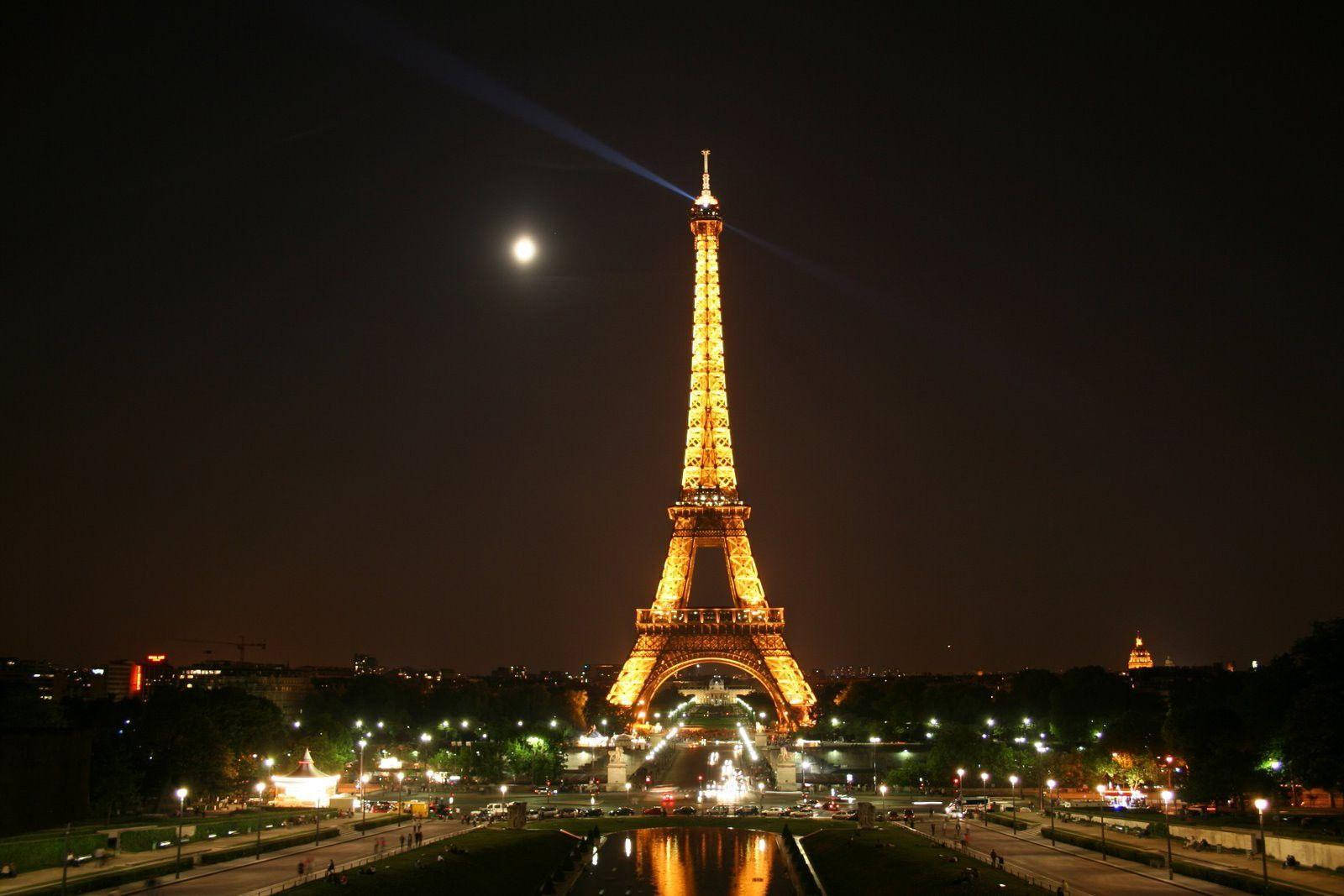Eiffel Tower 桌布 壁紙 — 艾菲爾鐵塔在明亮的月亮下溫暖地發光，光束從其頂部射向黑暗的巴黎夜空。