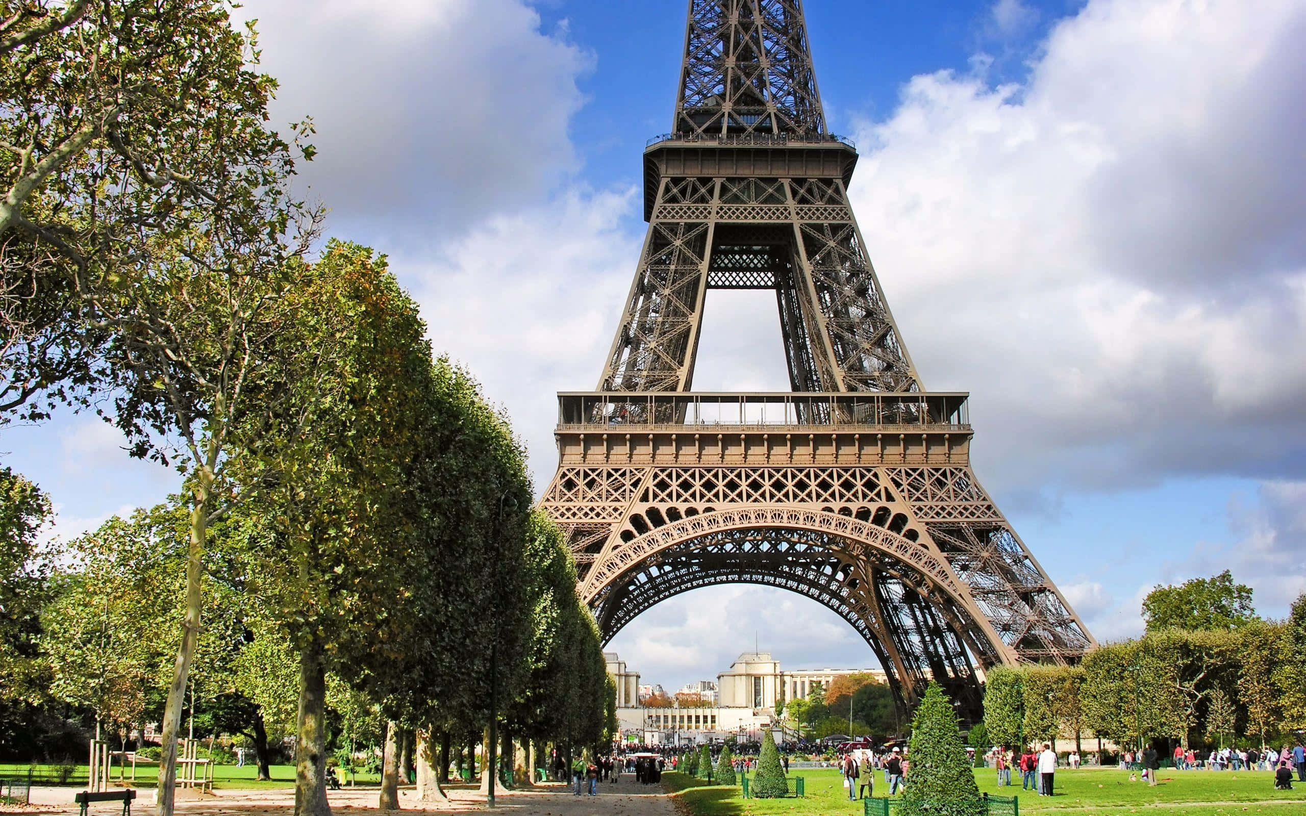 Eiffel Tower 桌布 壁紙 — 艾菲爾鐵塔在明亮的藍天和白雲下高聳，周圍環繞著茂密的綠樹和遊客熙攘的公園。