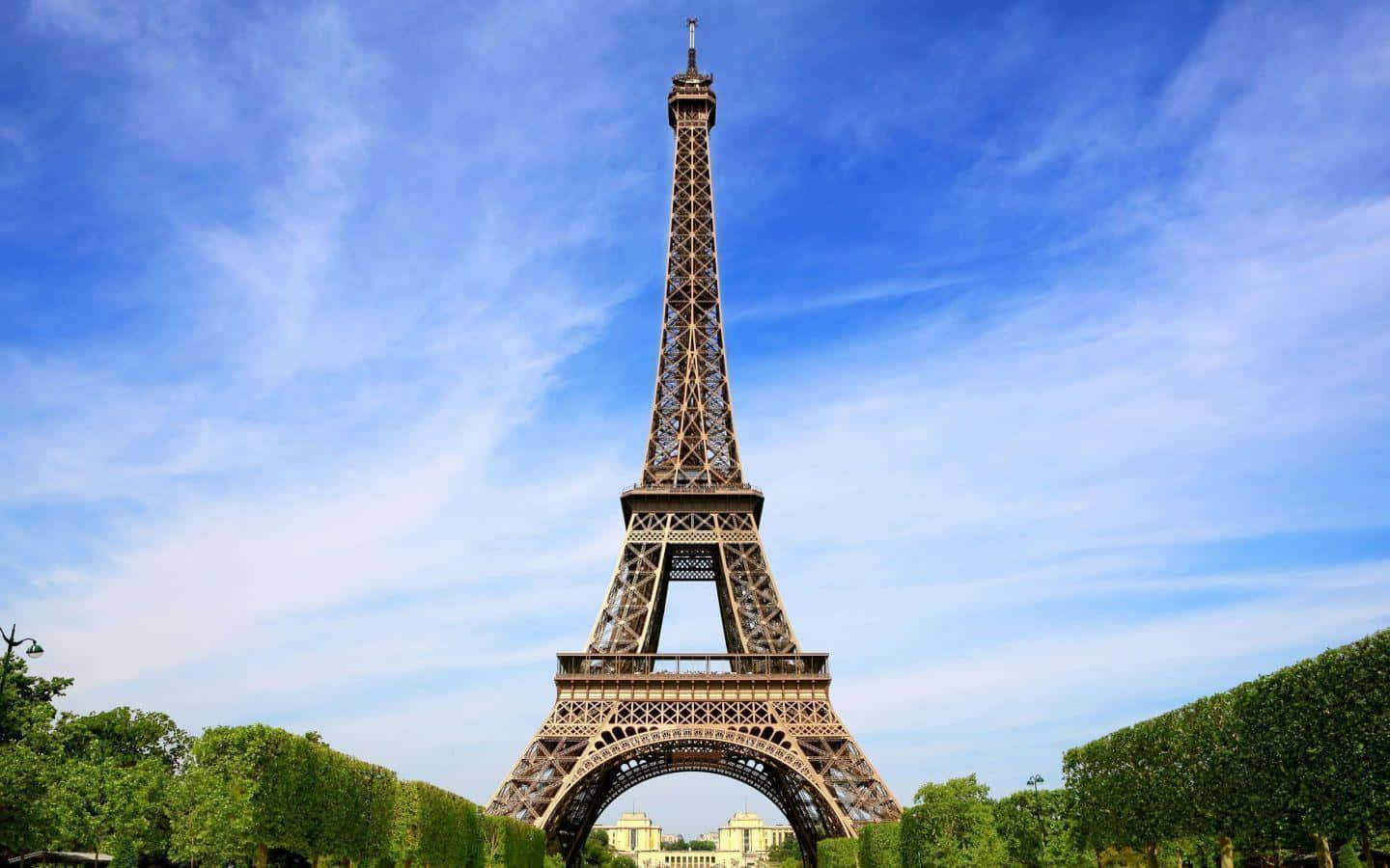 Eiffel Tower 桌布 壁紙 — 艾菲爾鐵塔在明亮的藍天和縷縷白雲襯托下高聳入雲，四周環繞著茂密的綠色樹籬。