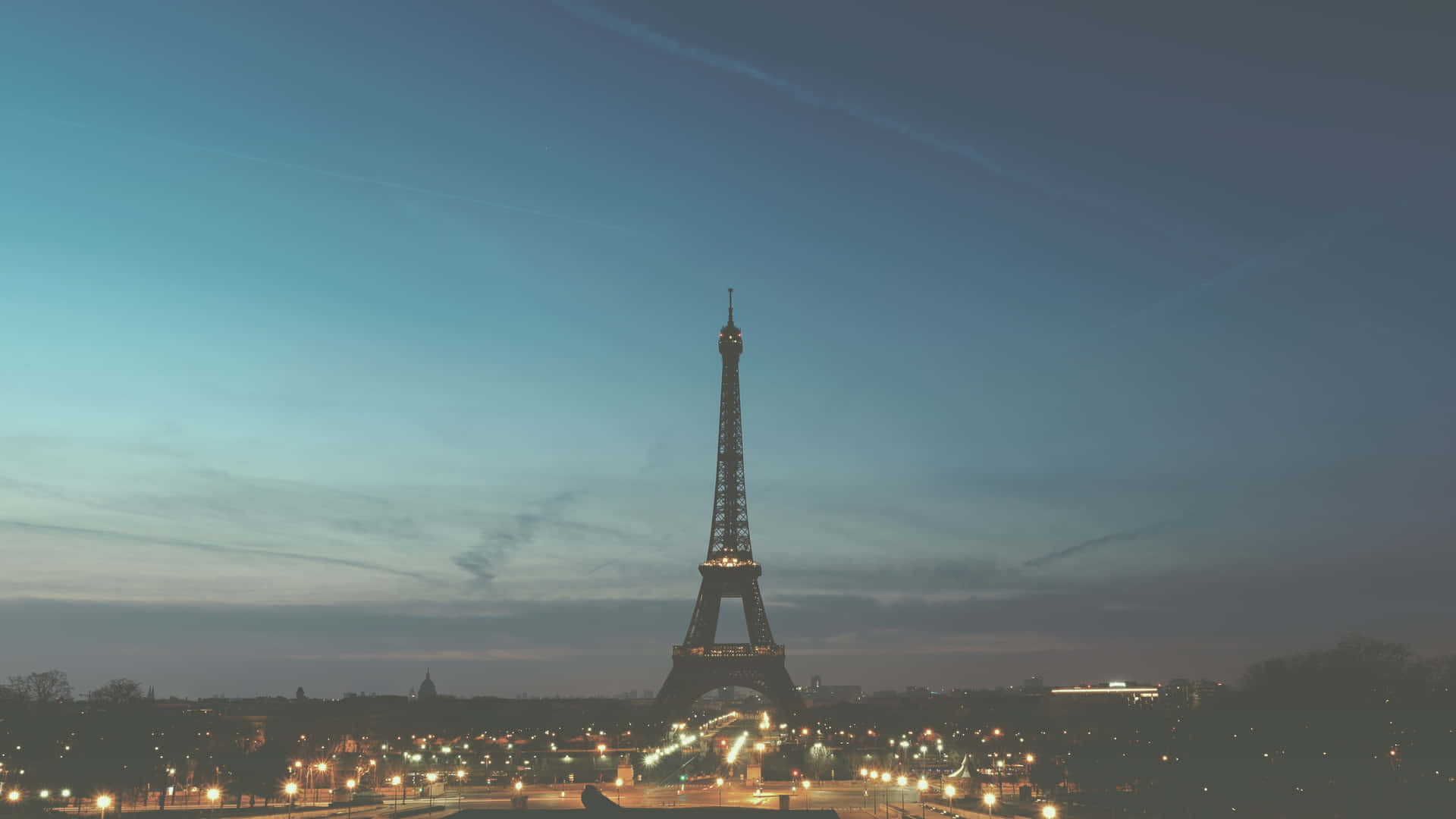 Eiffel Tower 桌布 壁紙 — 艾菲爾鐵塔在暮色天空下亮起，帶有柔和的藍色和灰色色調，下方散落著城市燈光。