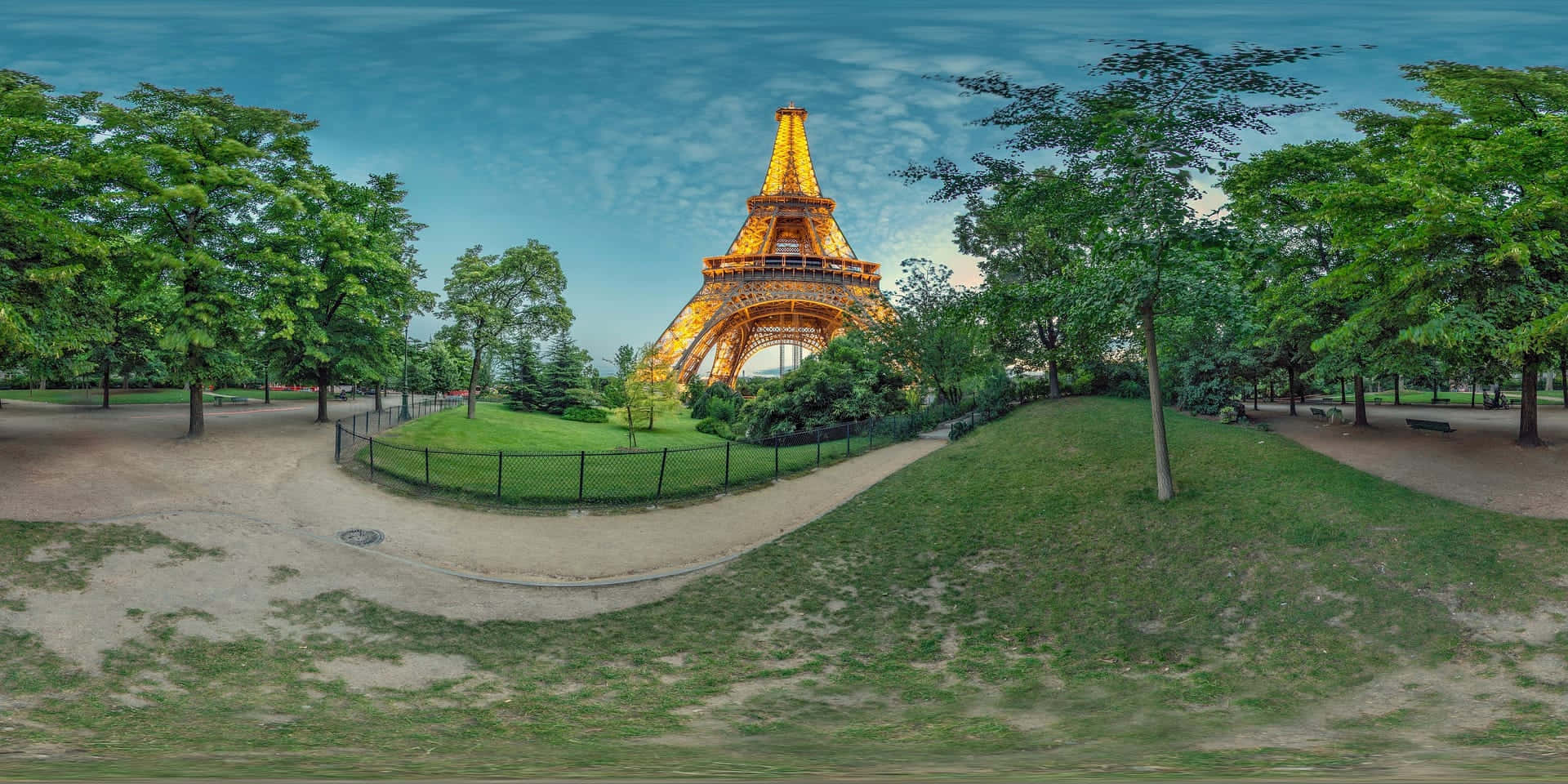 Eiffel Tower 桌布 壁紙 — 艾菲爾鐵塔在暮色天空下溫暖地發光，被茂密的綠樹和蜿蜒的公園小徑環繞。