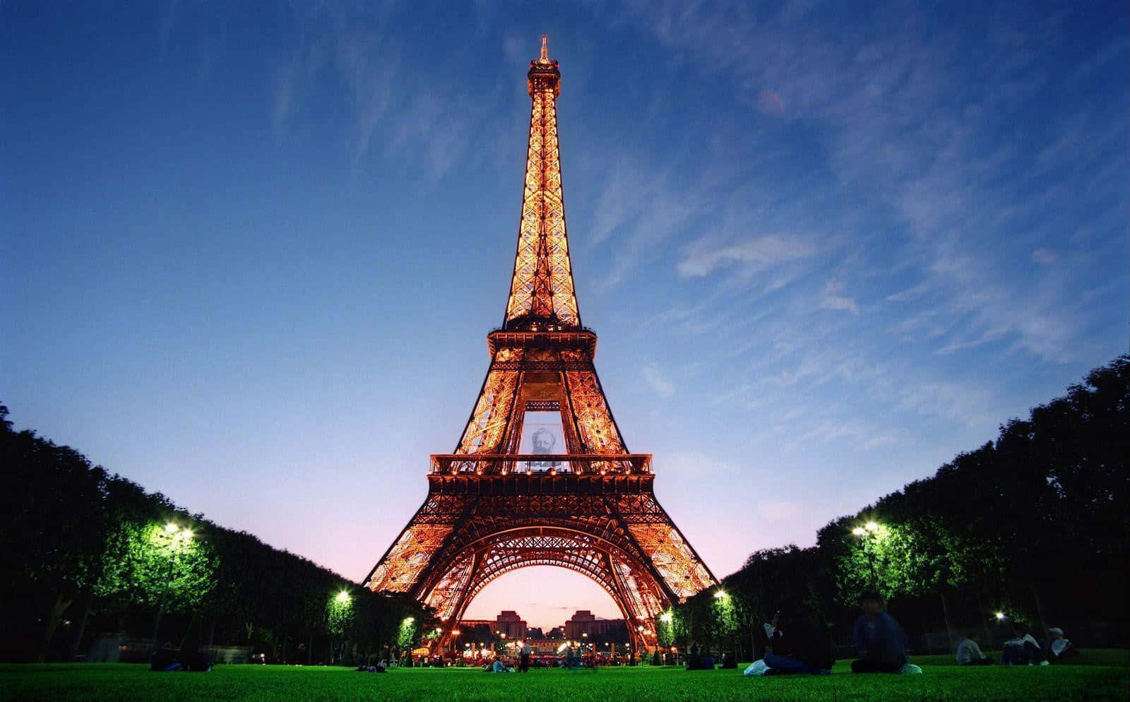 Eiffel Tower 桌布 壁紙 — 艾菲爾鐵塔在暮色天空下溫暖地發光，人們在下面的草地上放鬆。
