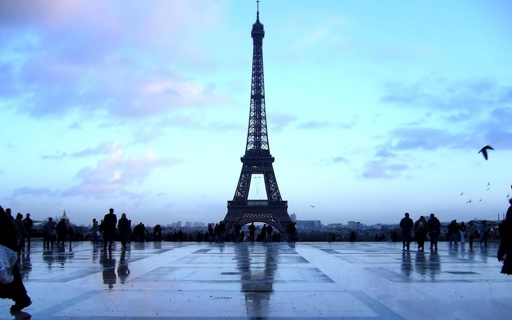 Eiffel Tower 桌布 壁紙 — 艾菲爾鐵塔在暮色天空下高聳，剪影人群聚集在潮濕反光的廣場上。