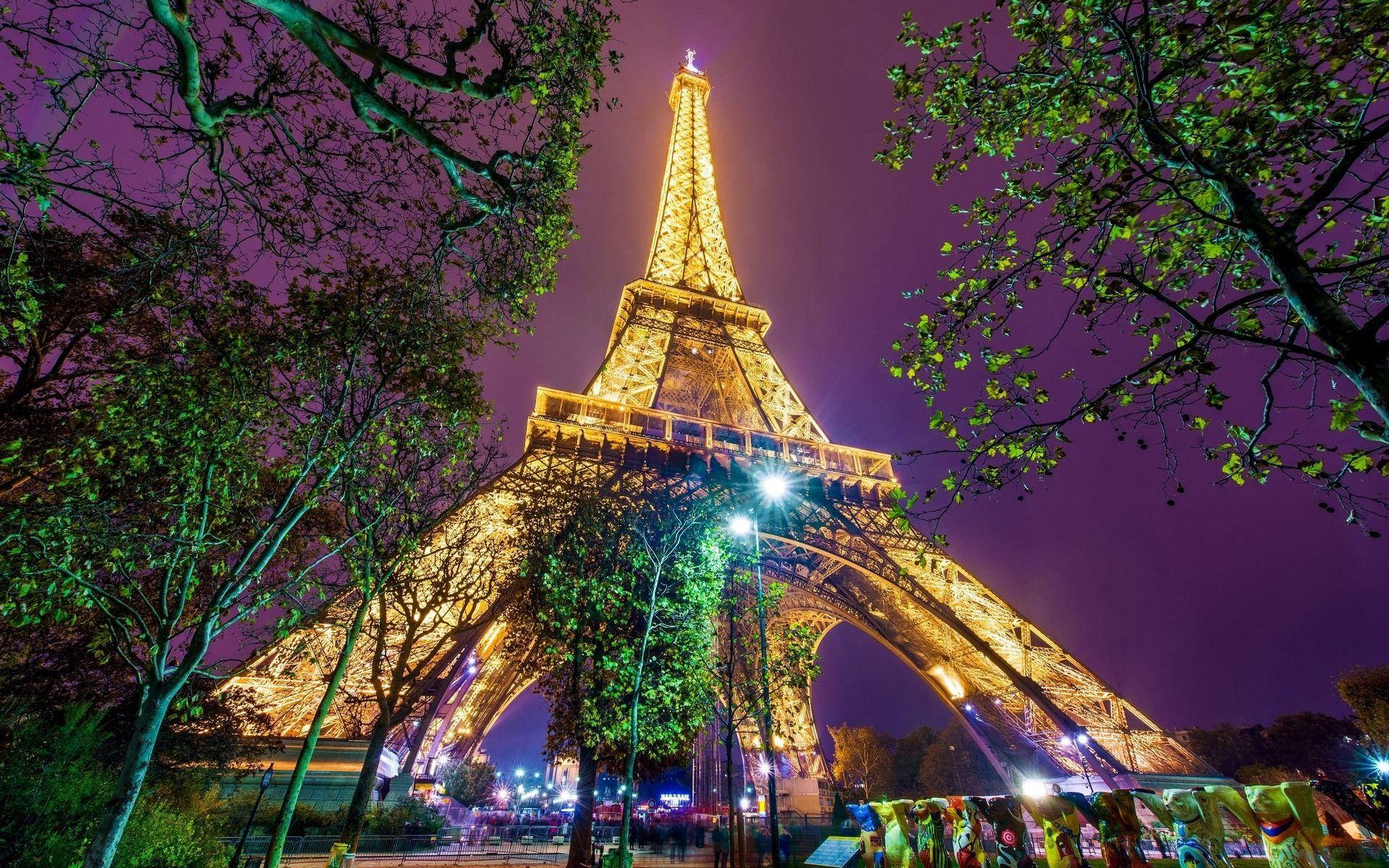 Paris Eiffel Tower 桌布 壁紙 — 艾菲爾鐵塔在深紫色的夜空下溫暖地發光，周圍是長出嫩芽的樹木剪影。