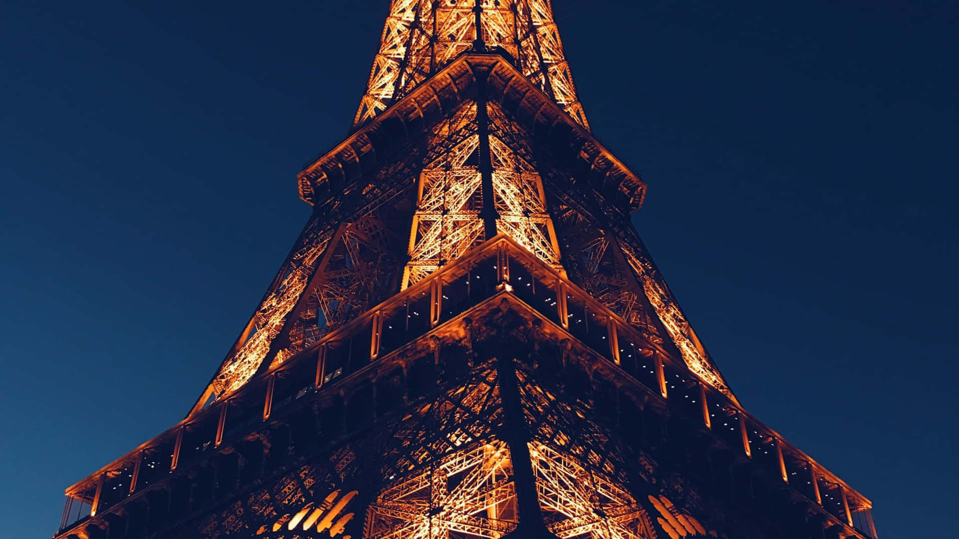 Eiffel Tower 桌布 壁紙 — 艾菲爾鐵塔在深藍色夜空中溫暖發光，以戲劇性的低角度拍攝。