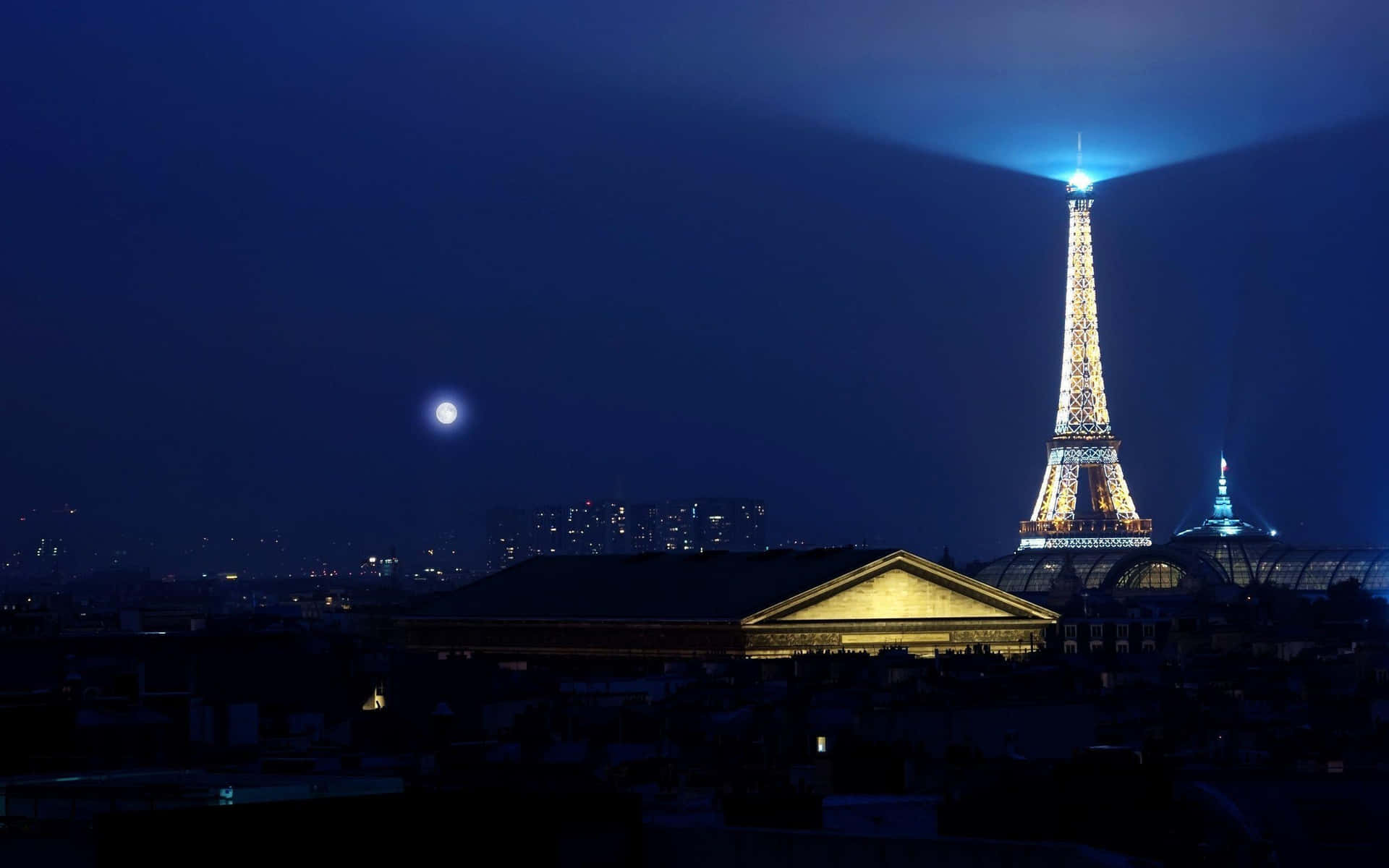 Eiffel Tower 桌布 壁紙 — 艾菲爾鐵塔在深藍色的夜空中溫暖地發光，附近有一輪明亮的月亮，下方城市燈光閃爍。
