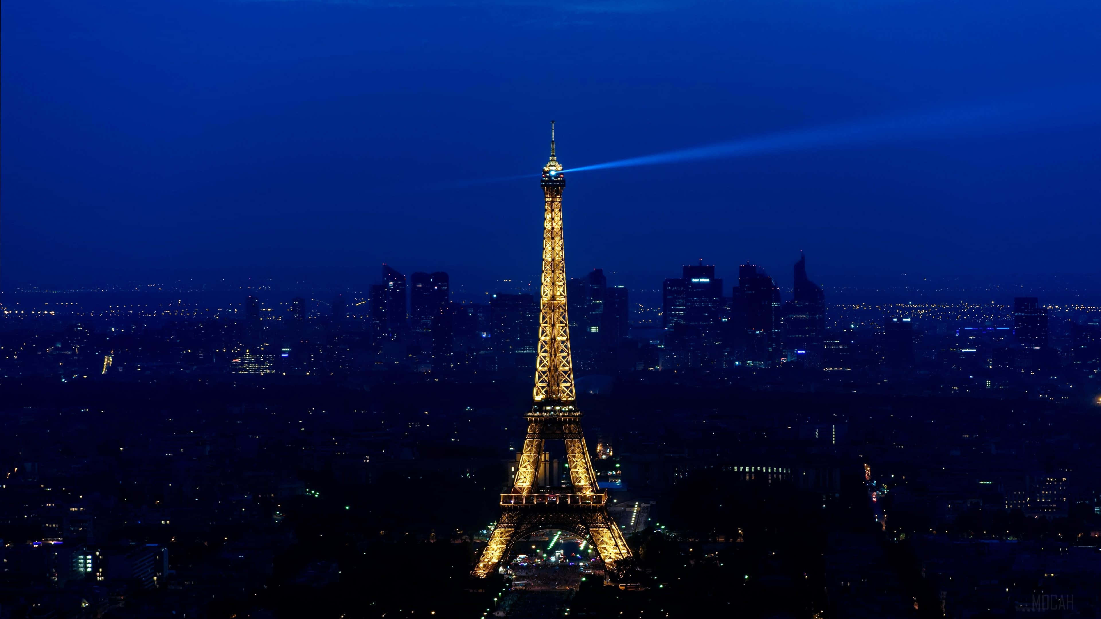 Paris At Night 桌布 壁紙 — 艾菲爾鐵塔在深藍色的夜空中溫暖地發光，並將聚光燈照射在巴黎燈火通明的天際線上。