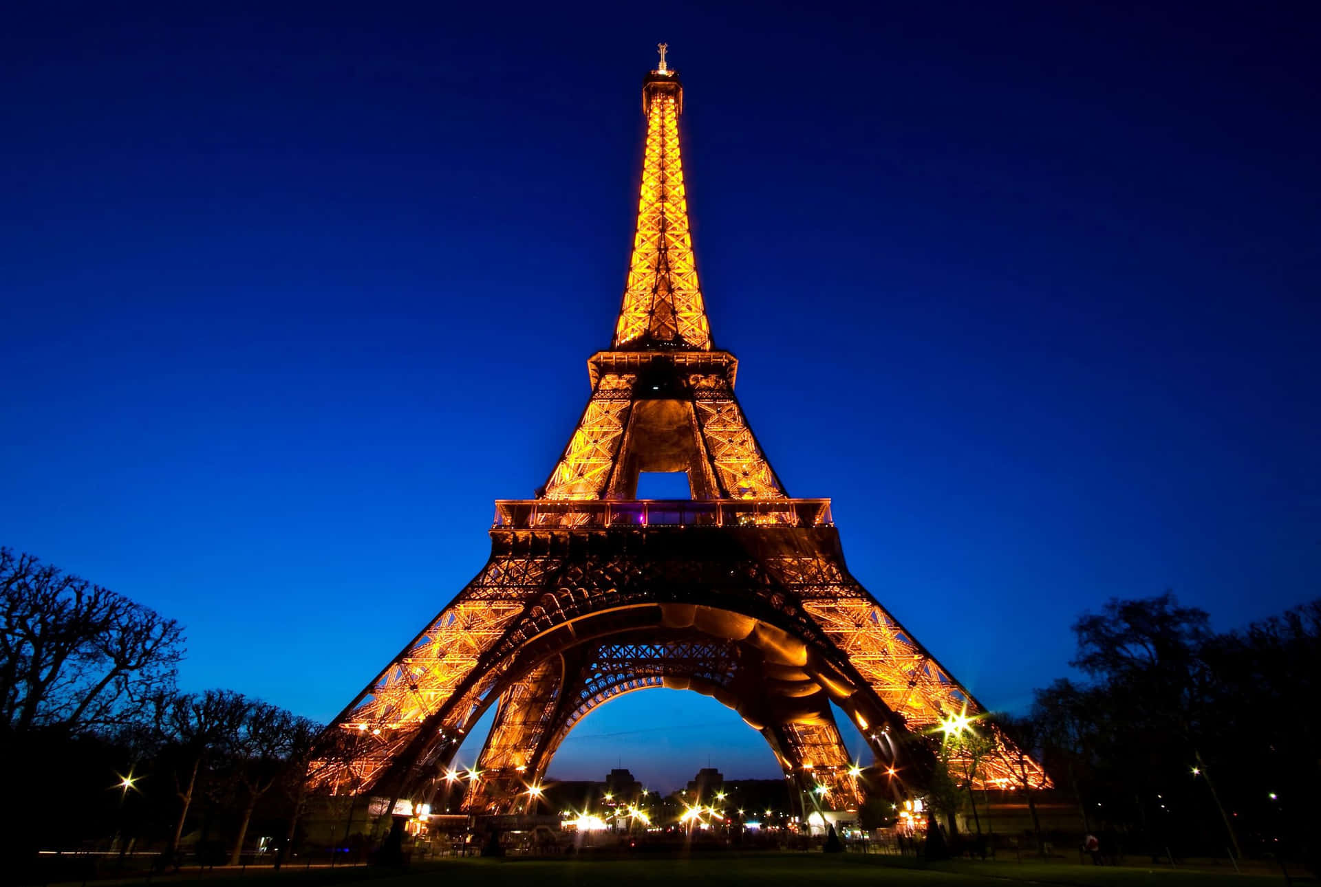 Eiffel Tower 桌布 壁紙 — 艾菲爾鐵塔在深藍色的暮色天空中溫暖地發光，剪影般的樹木襯托出其雄偉的鐵結構。